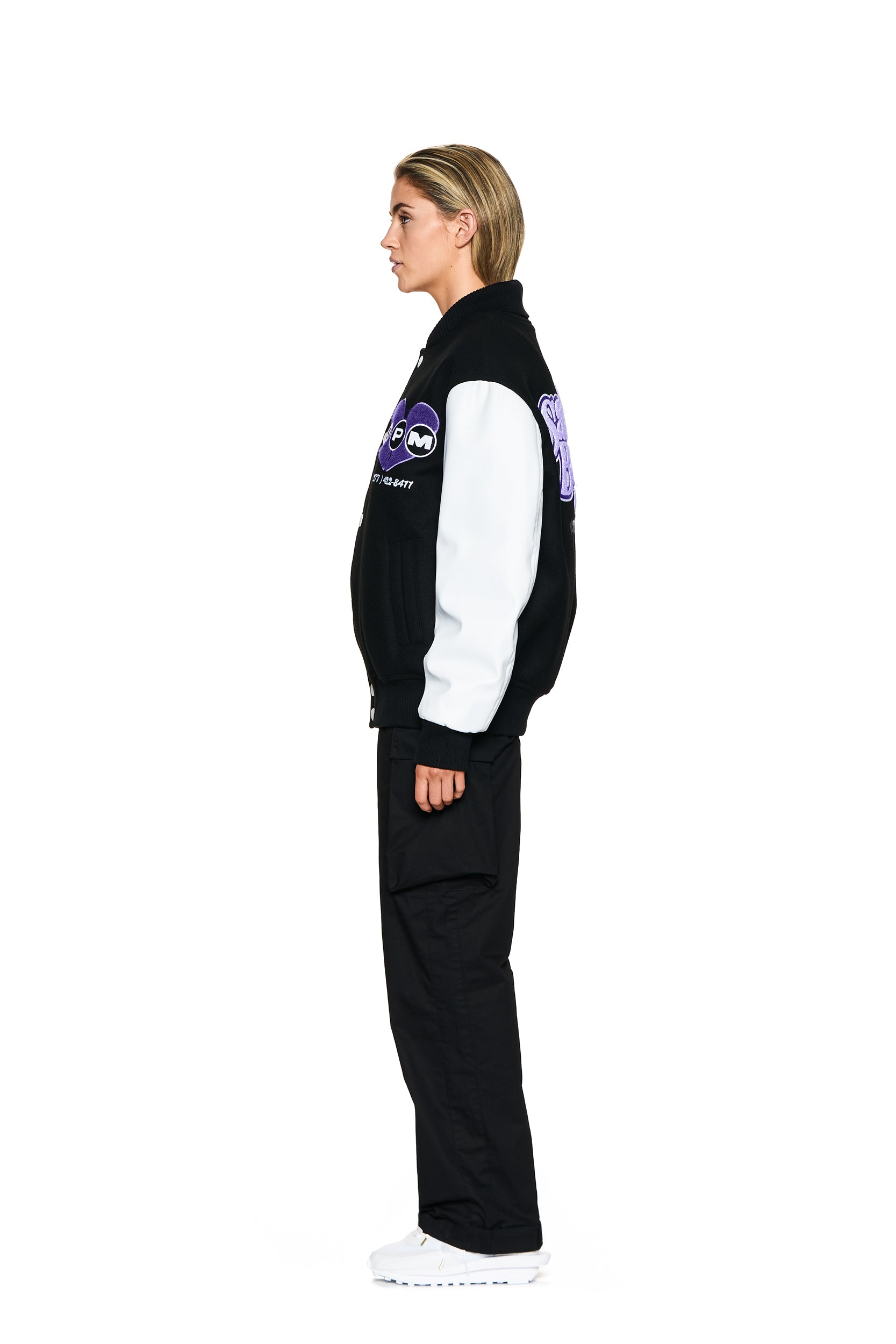 A person with short blonde hair stands in profile, wearing the 6PM COLLEGE JACKET BLACK, black pants, and white sneakers against a plain white background.