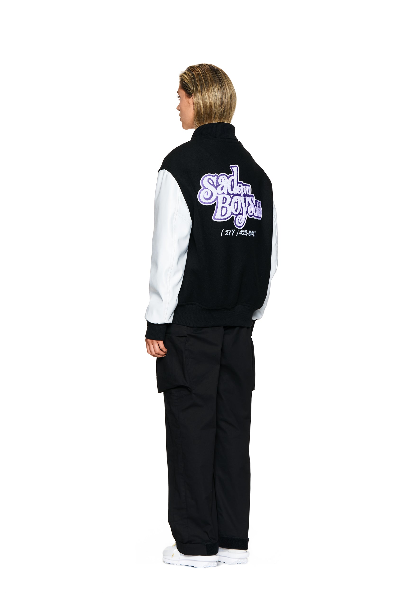 A person stands facing away, wearing the 6PM COLLEGE JACKET BLACK varsity jacket with purple and white back graphics, black cargo pants, and white sneakers, set against a plain white background.