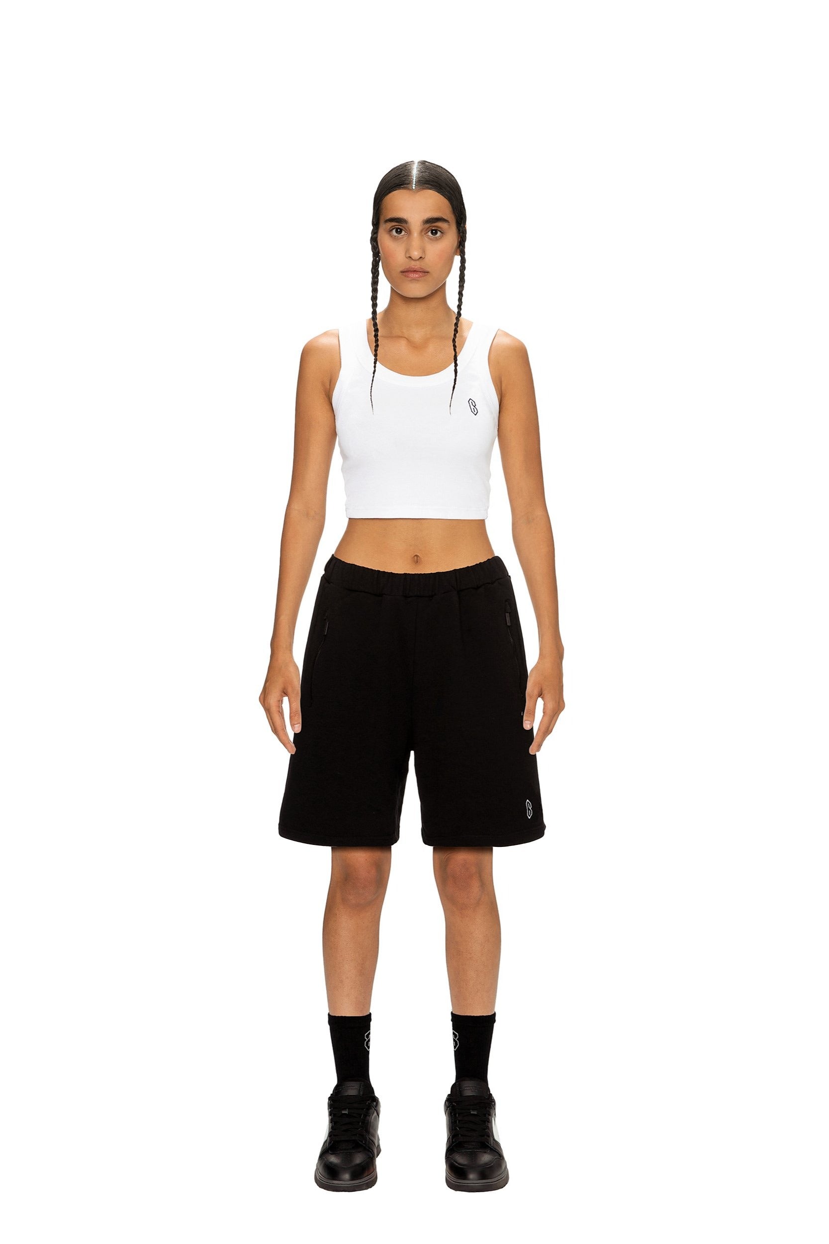 A person with braided hair faces forward against a white background, wearing the 6PM CROP TOP (2-PACK) in white, paired with black knee-length shorts, black socks, and black shoes.