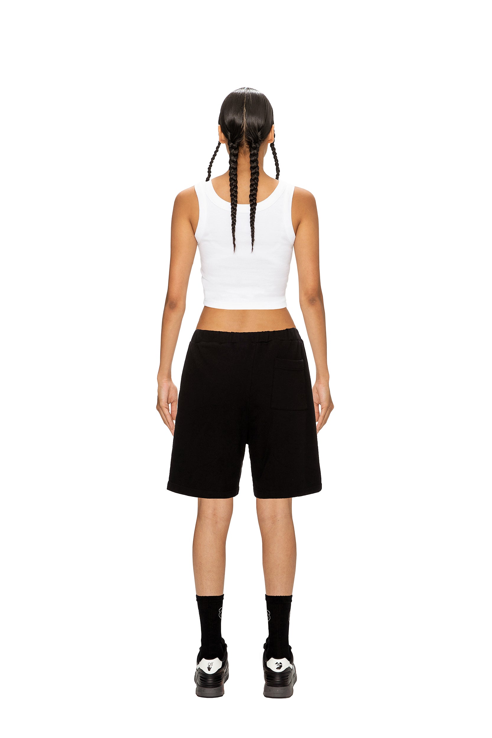 A person with two long braids, wearing a white 6PM CROP TOP (2-PACK), black knee-length shorts, black socks, and black shoes stands facing away against a plain white background.