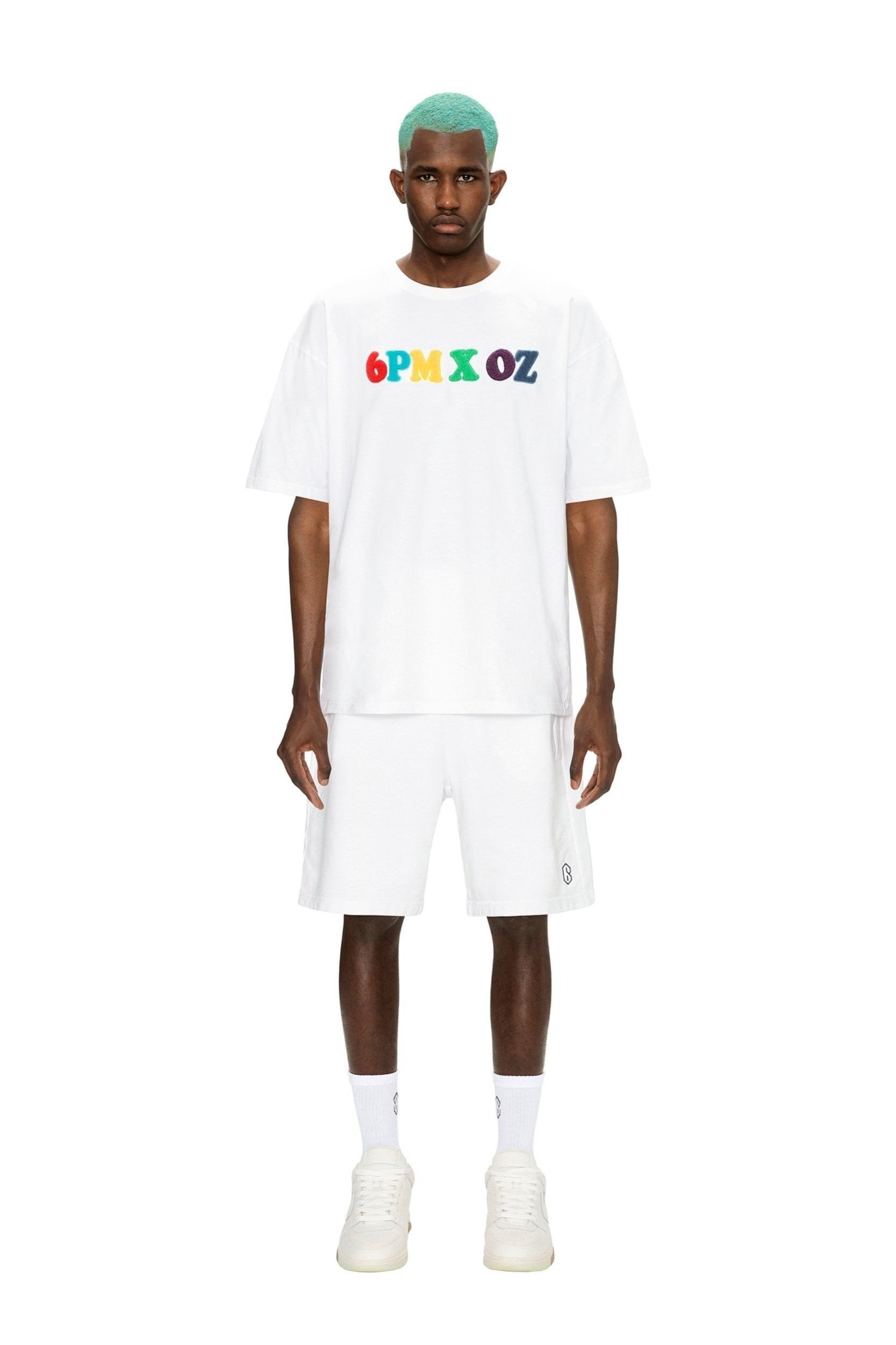 A man with turquoise hair wears the 6PM RAINBOW T-SHIRT WHITE, white shorts, white socks, and white sneakers, standing against a plain white background.