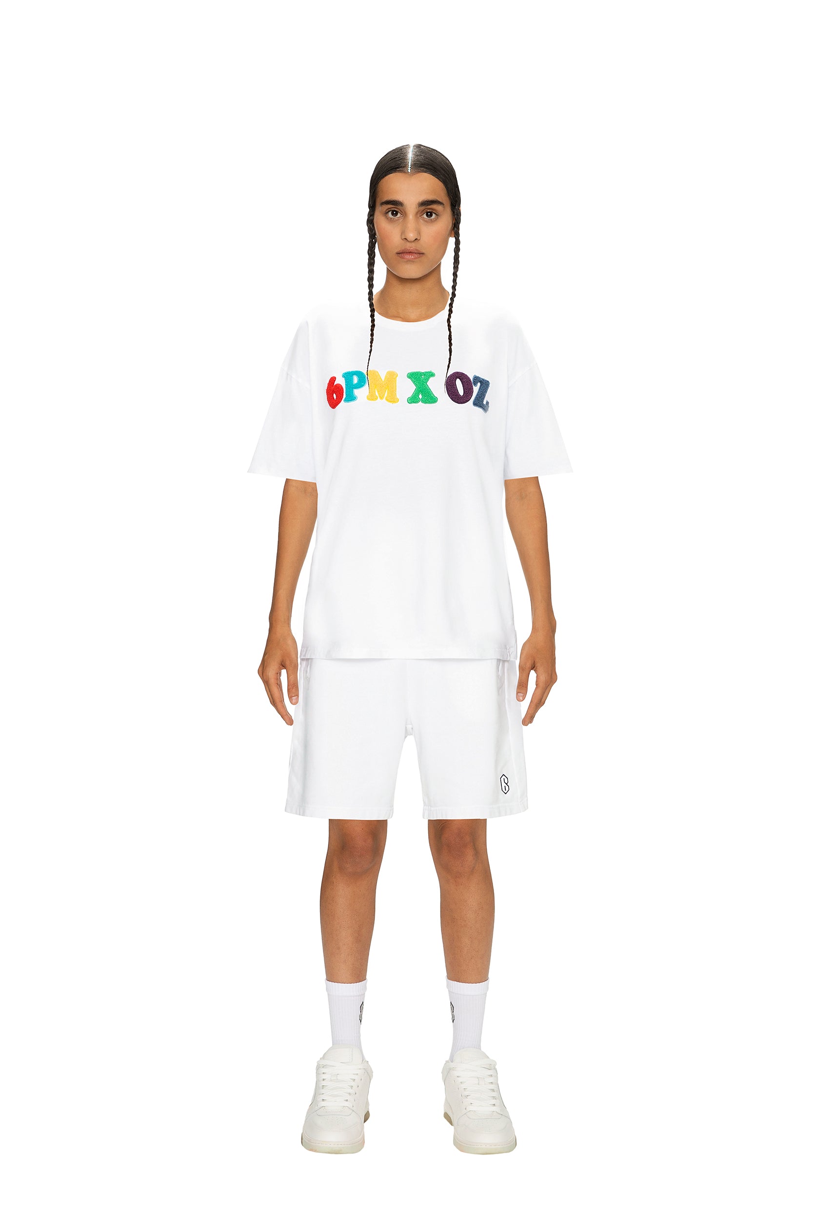 A person with braided hair faces forward, wearing the 6PM RAINBOW T-SHIRT WHITE, matching white shorts, socks, and sneakers, all set against a plain white background.