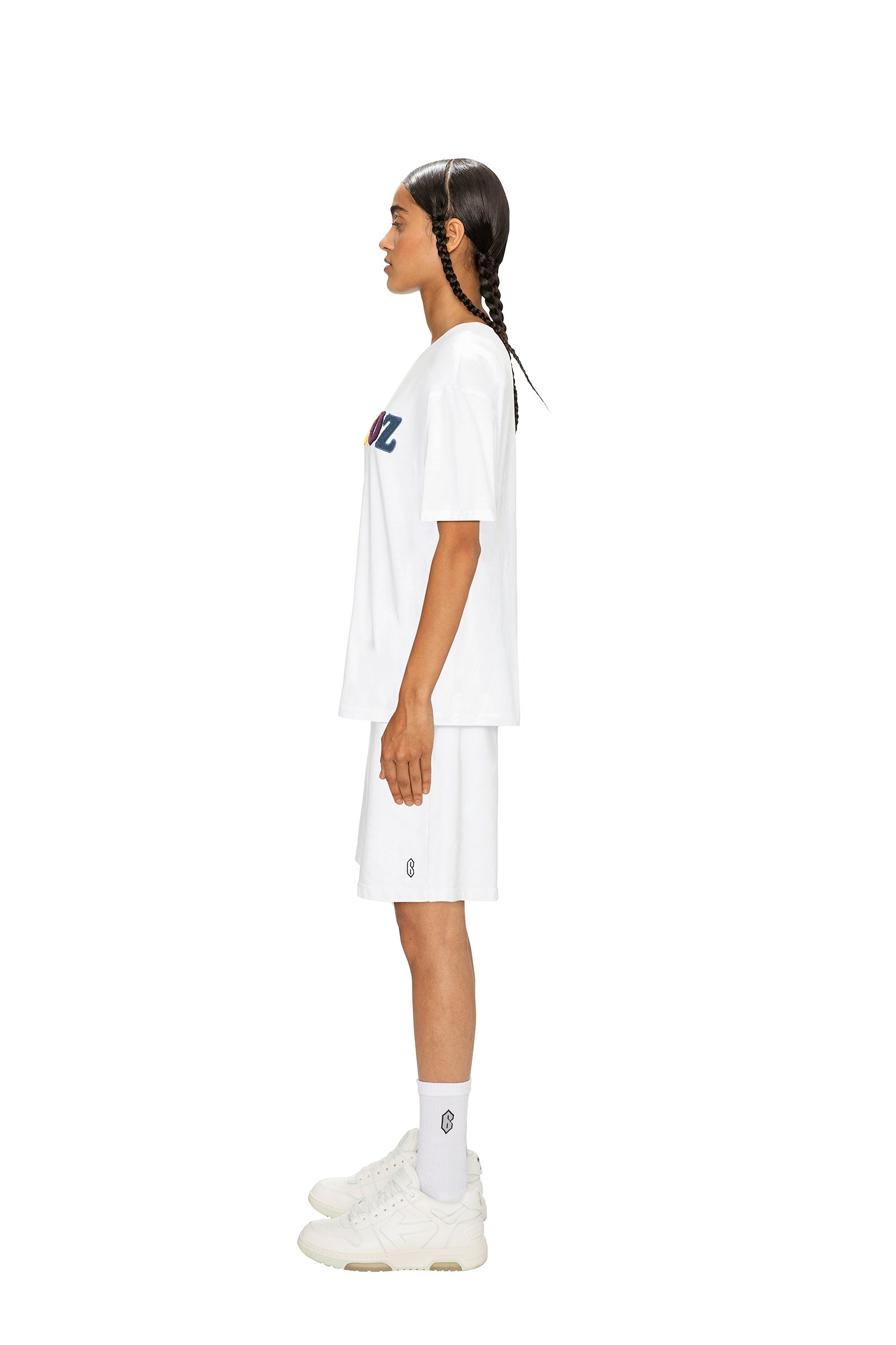 A person with long braids stands in profile, wearing the 6PM RAINBOW T-SHIRT WHITE, white shorts, socks, and sneakers against a plain white background.