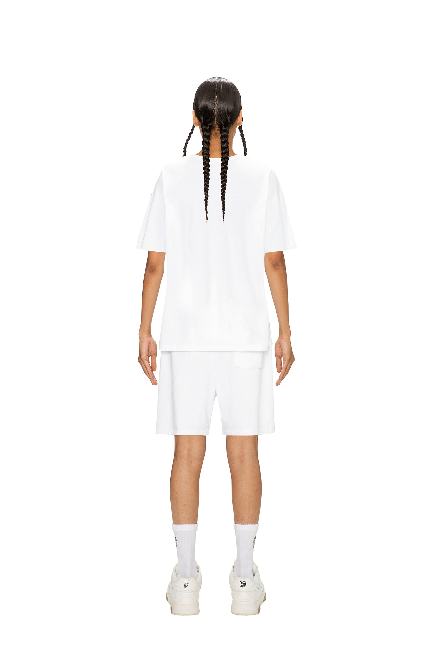 A person with long braids faces away, wearing the 6PM RAINBOW T-SHIRT WHITE, white shorts, socks, and sneakers, all set against a plain white background.