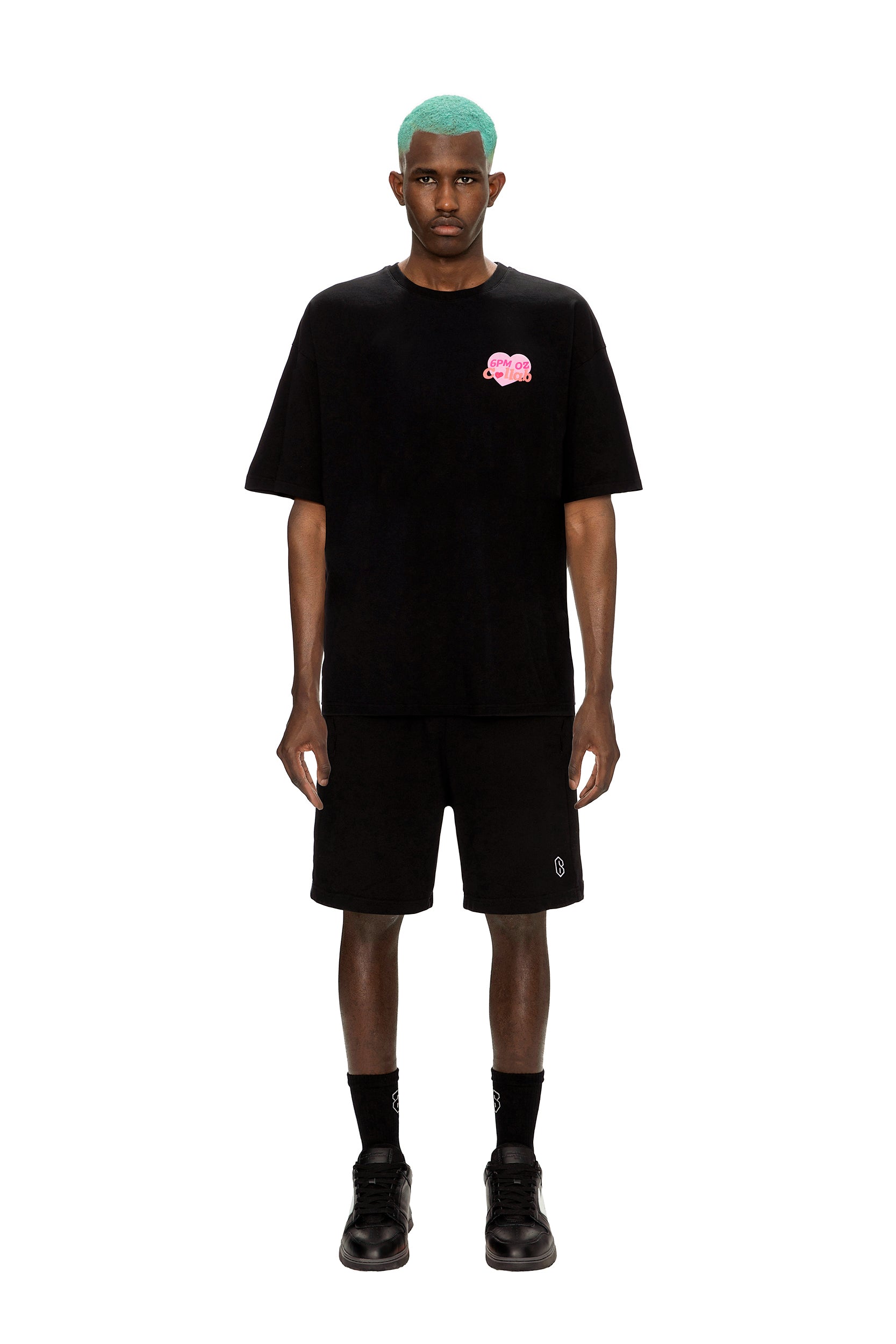 A man with turquoise hair wears the 6PM COLLAB T-SHIRT BLACK featuring a small pink graphic, paired with black shorts, socks, and shoes, standing against a white background.