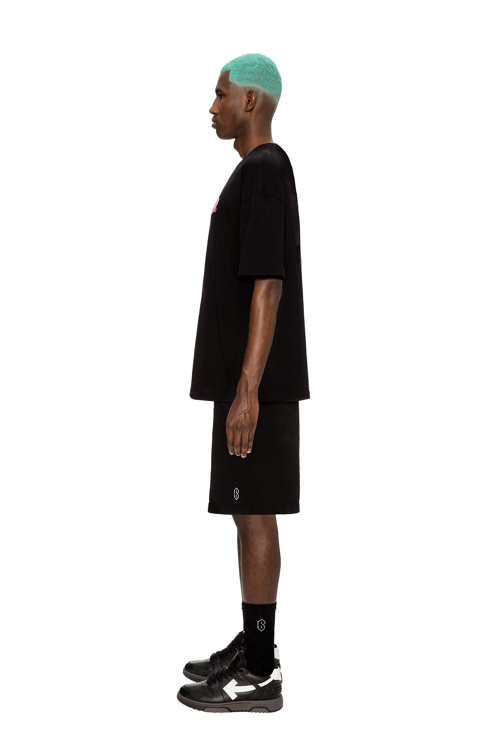 A man with teal hair stands in profile against a white background, wearing the 6PM COLLAB T-SHIRT BLACK, paired with black shorts, socks, and sneakers.