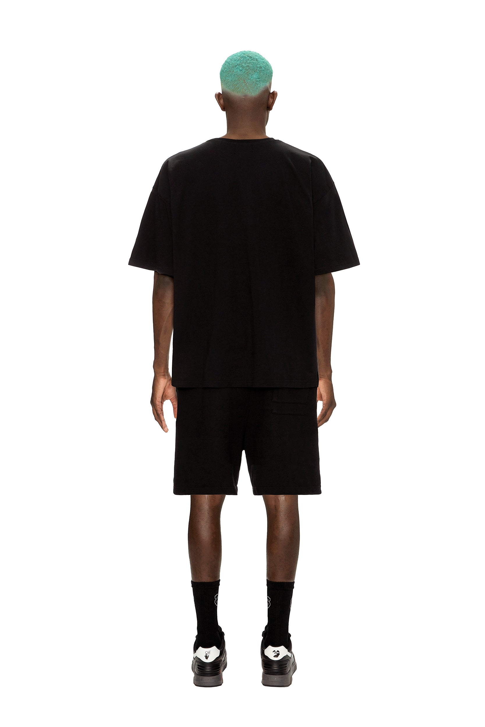 A person with short turquoise hair stands facing away, wearing the 6PM COLLAB T-SHIRT BLACK along with black shorts, socks, and shoes against a plain white background.