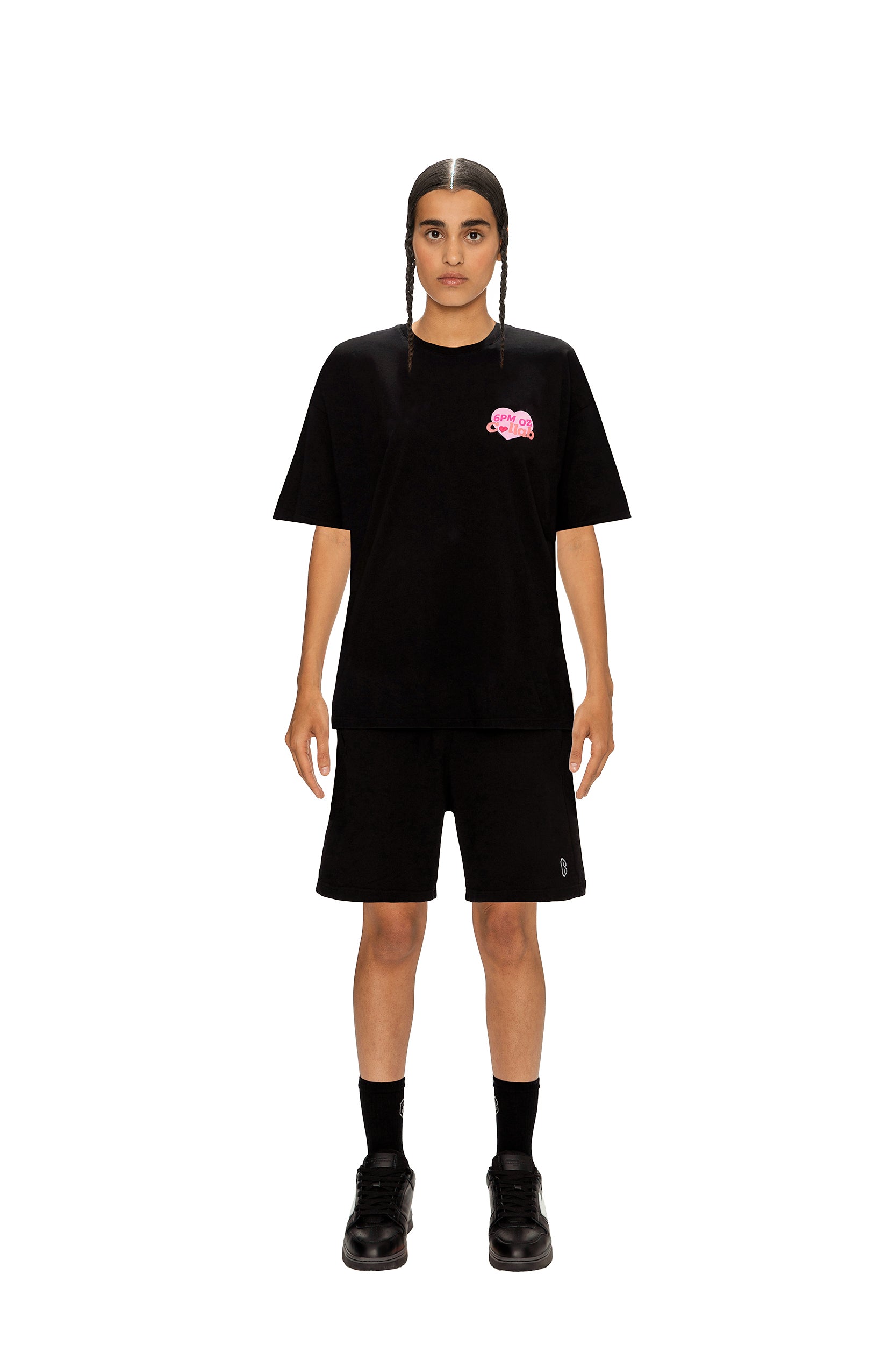 A person with two long braids stands against a white background wearing the 6PM COLLAB T-SHIRT BLACK, featuring a pink chest graphic, paired with black shorts, socks, and shoes.
