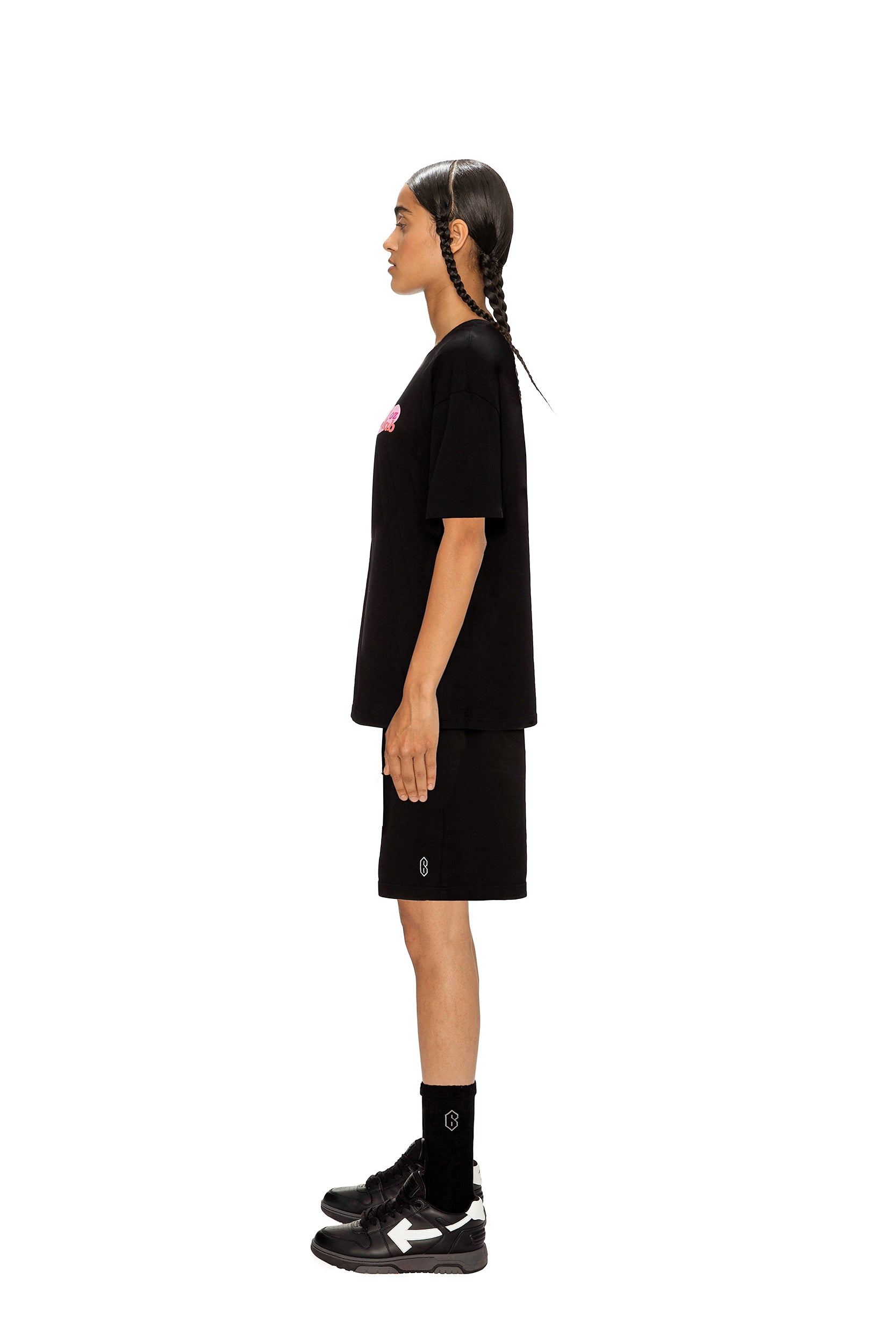 A person with two braids stands in profile facing left, wearing the 6PM COLLAB T-SHIRT BLACK, black shorts, black socks, and black sneakers against a plain white background.