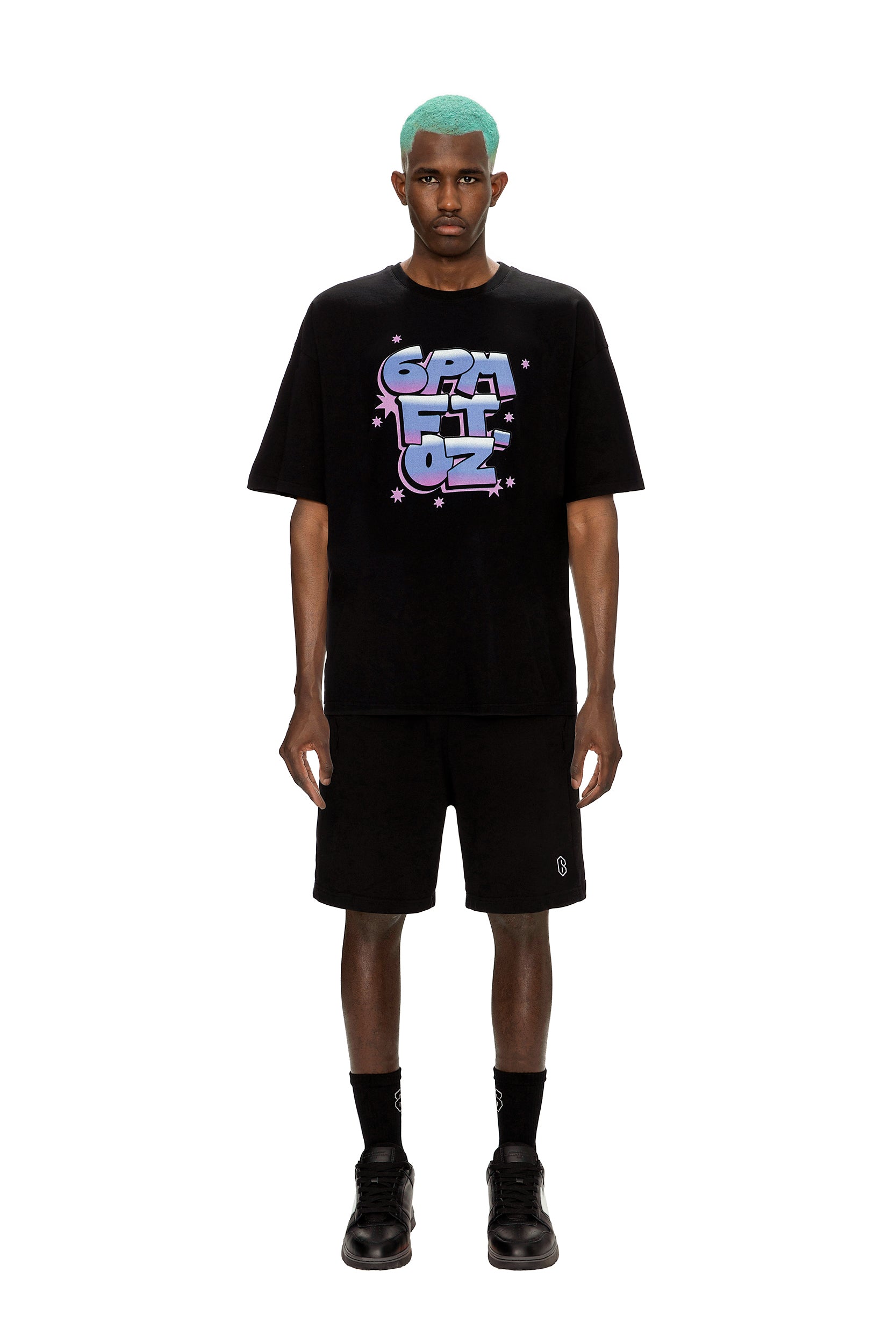 A man with teal hair stands facing forward in a 6PM AIRBRUSH T-SHIRT BLACK, black shorts, socks, and shoes against a plain white background.