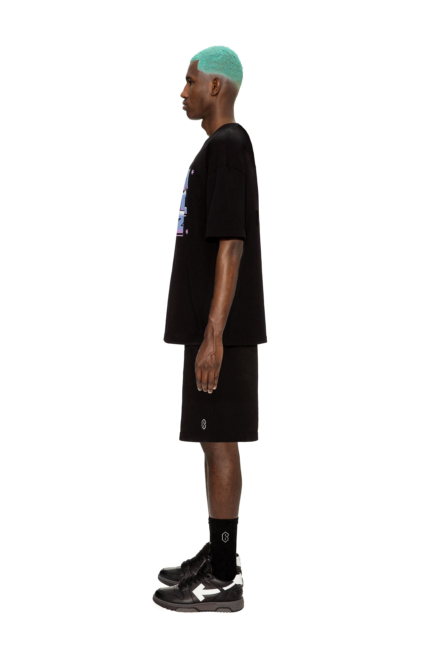 A man with turquoise hair stands in profile against a white background, wearing the 6PM AIRBRUSH T-SHIRT BLACK, black shorts, black socks, and black sneakers.