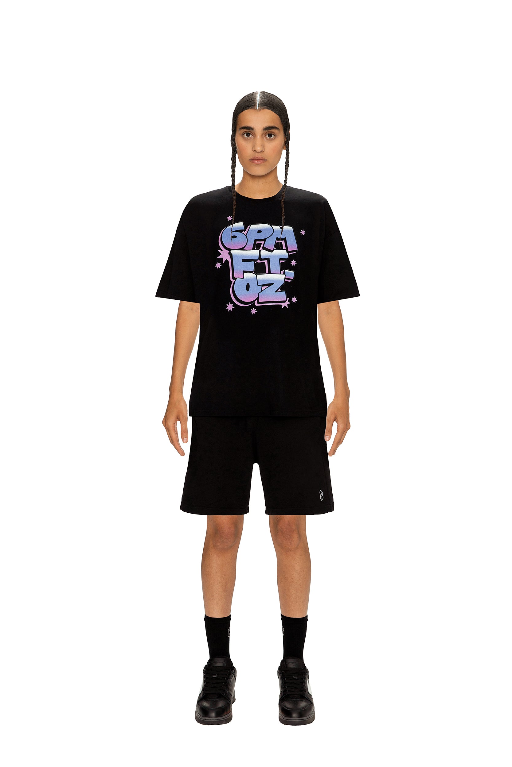 A person with braided hair stands facing forward, wearing the 6PM AIRBRUSH T-SHIRT BLACK with pastel SPACFLOZ text and star graphics, paired with black shorts, socks, and shoes against a white background.