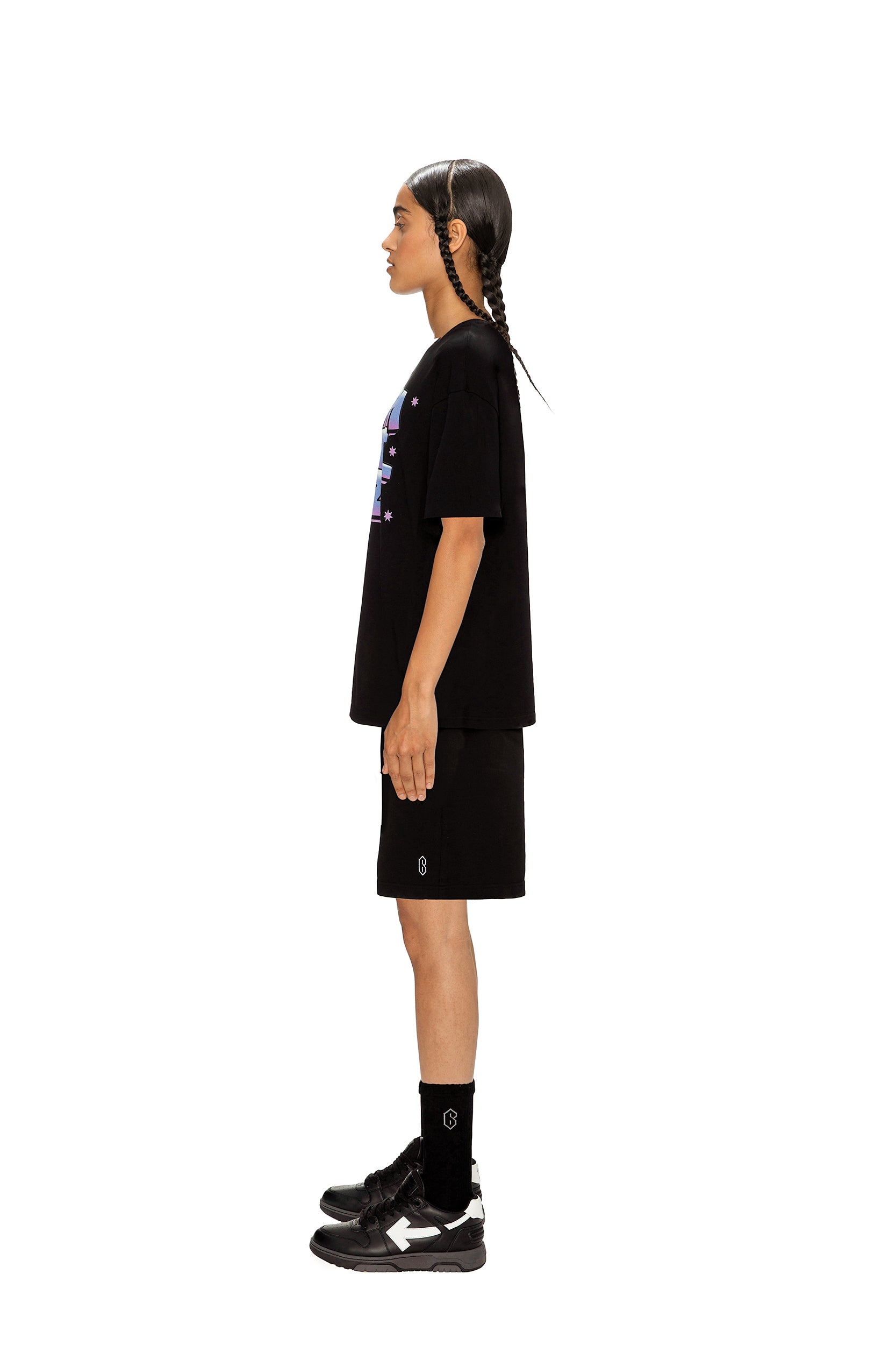 Person with braided hair stands in profile against a white background, wearing the 6PM AIRBRUSH T-SHIRT BLACK, black shorts, black socks, and black sneakers.