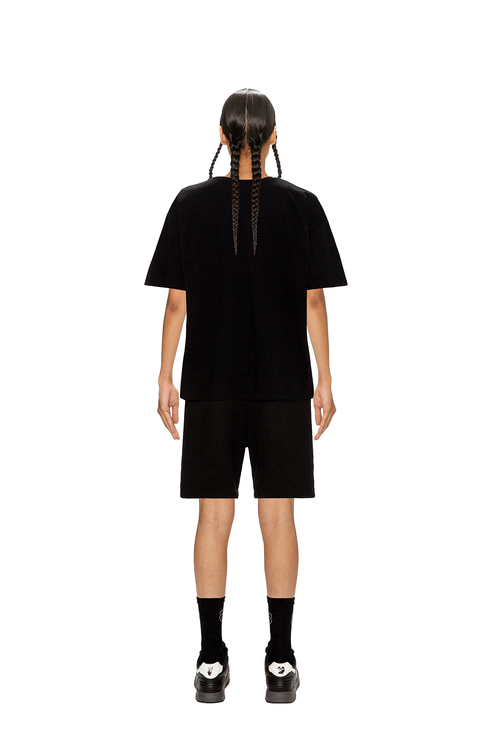 A person with two long braids stands with their back to the camera, wearing the 6PM AIRBRUSH T-SHIRT BLACK, black shorts, socks, and shoes against a white background.