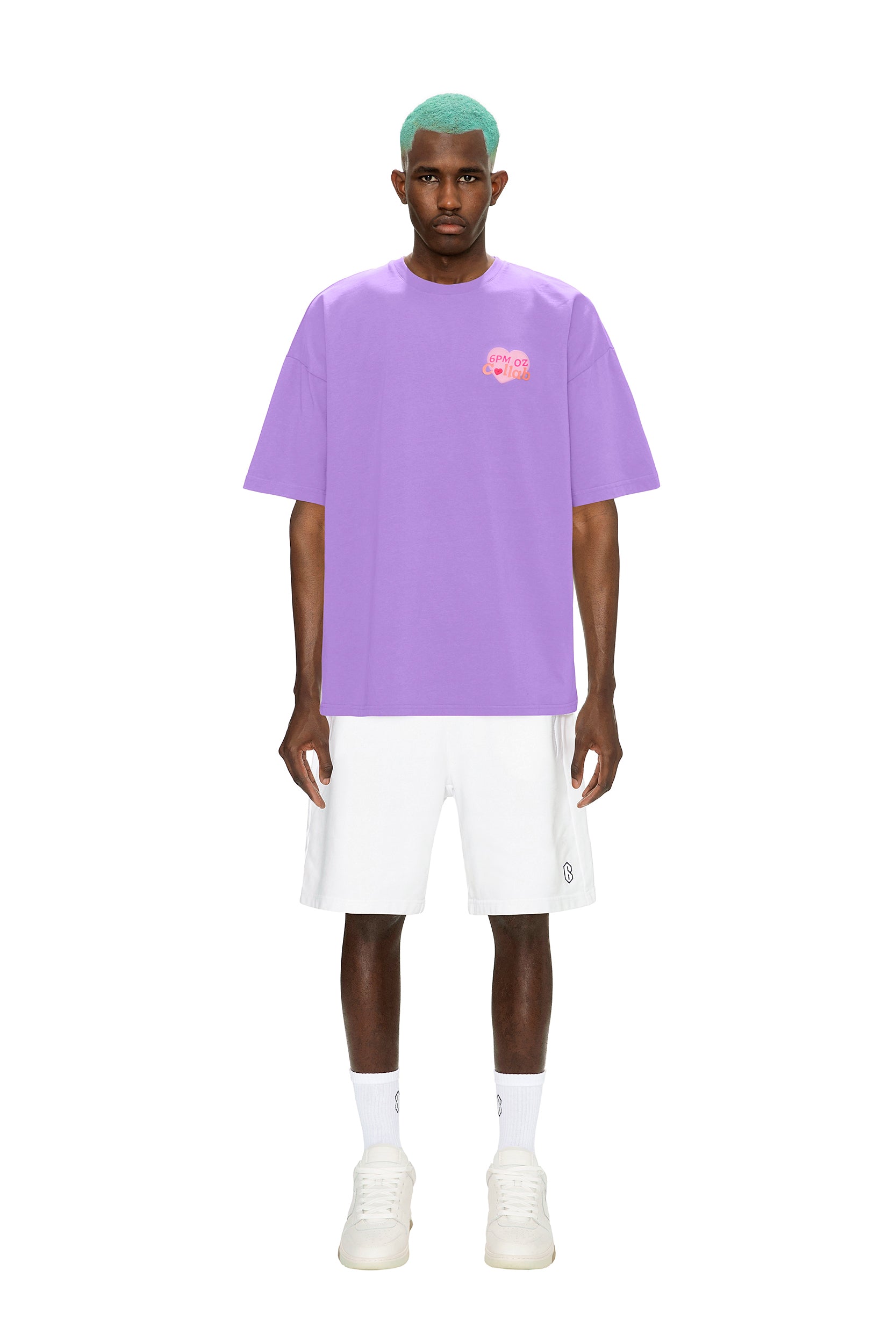 A man with teal hair faces forward against a white background, wearing the 6PM COLLAB T-SHIRT LILAC featuring a small pink chest graphic, paired with white shorts, socks, and sneakers.