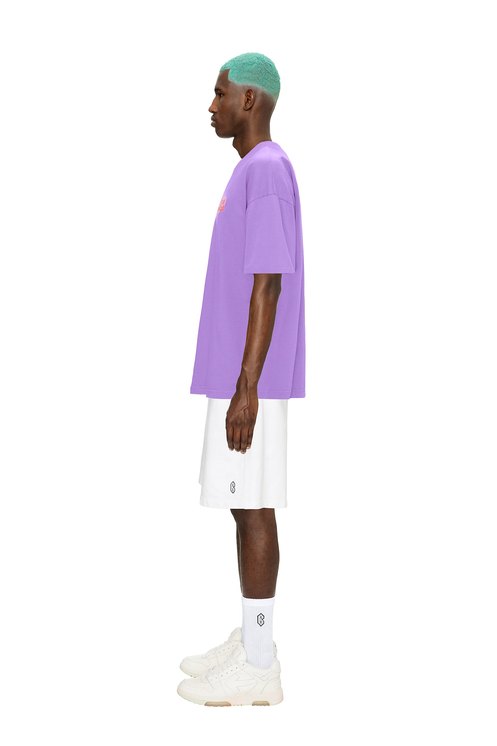 A man with teal hair stands in profile against a plain white background, wearing the 6PM COLLAB T-SHIRT LILAC paired with white shorts, white crew socks, and white sneakers.
