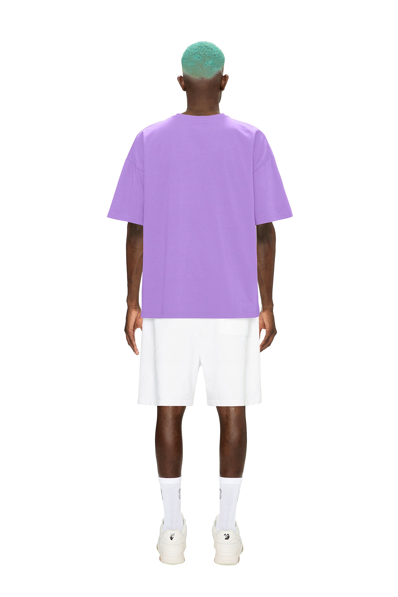 A person with teal hair stands facing away against a white background, wearing the 6PM COLLAB T-SHIRT LILAC, paired with white shorts, socks, and sneakers.