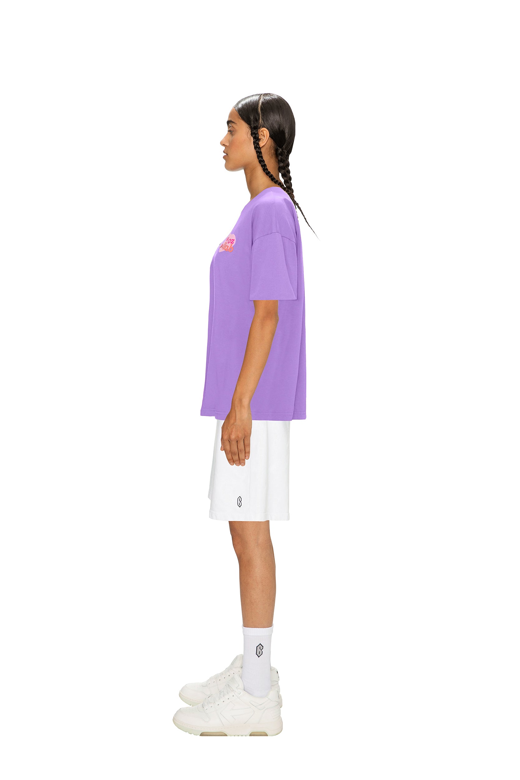 A person with two braids stands in profile against a white background, wearing the 6PM COLLAB T-SHIRT LILAC, white shorts, white socks, and white sneakers.