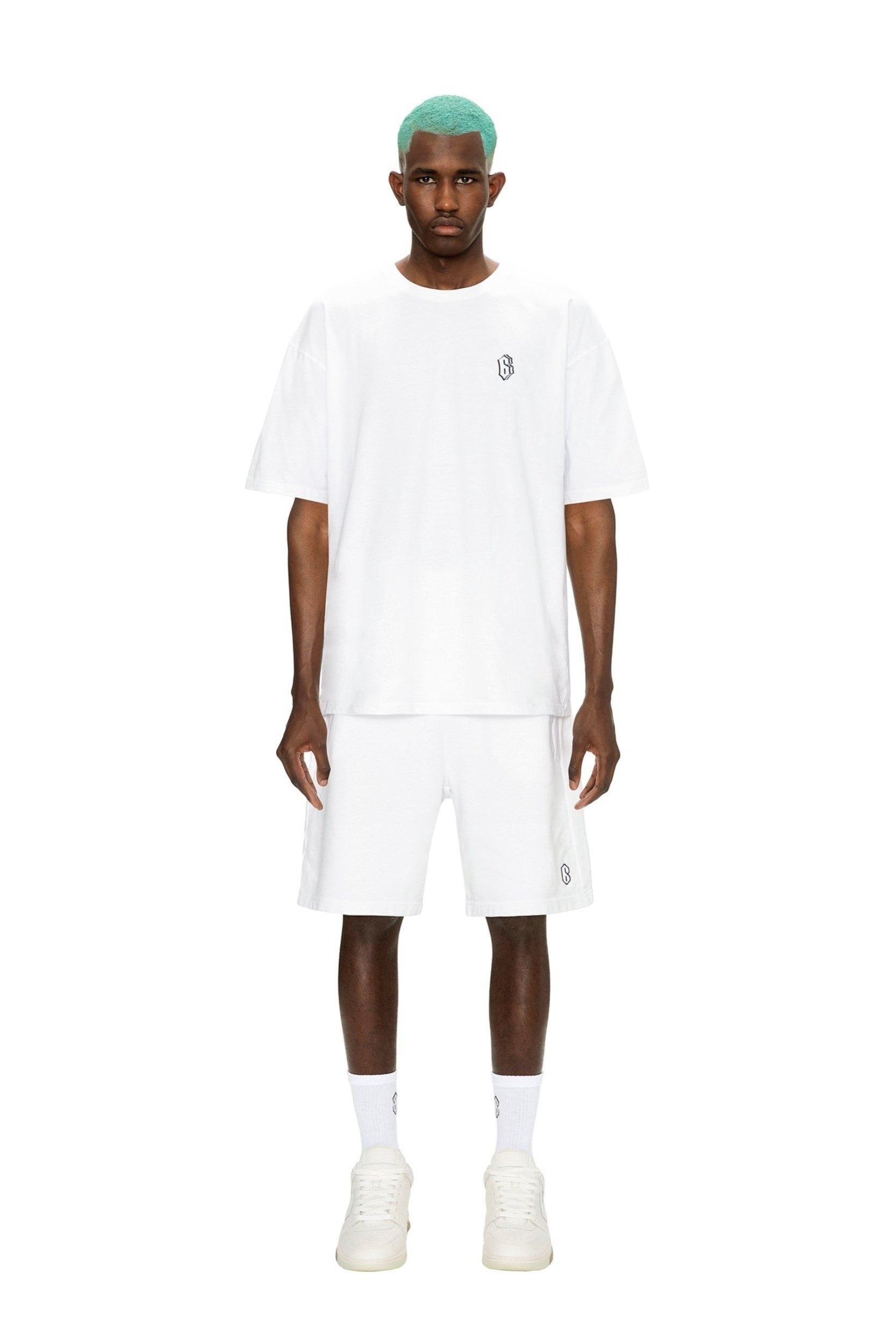A man with mint green hair wears the 6PM SIGNATURE T-SHIRT WHITE, paired with white shorts, socks, and sneakers, standing against a plain white background.