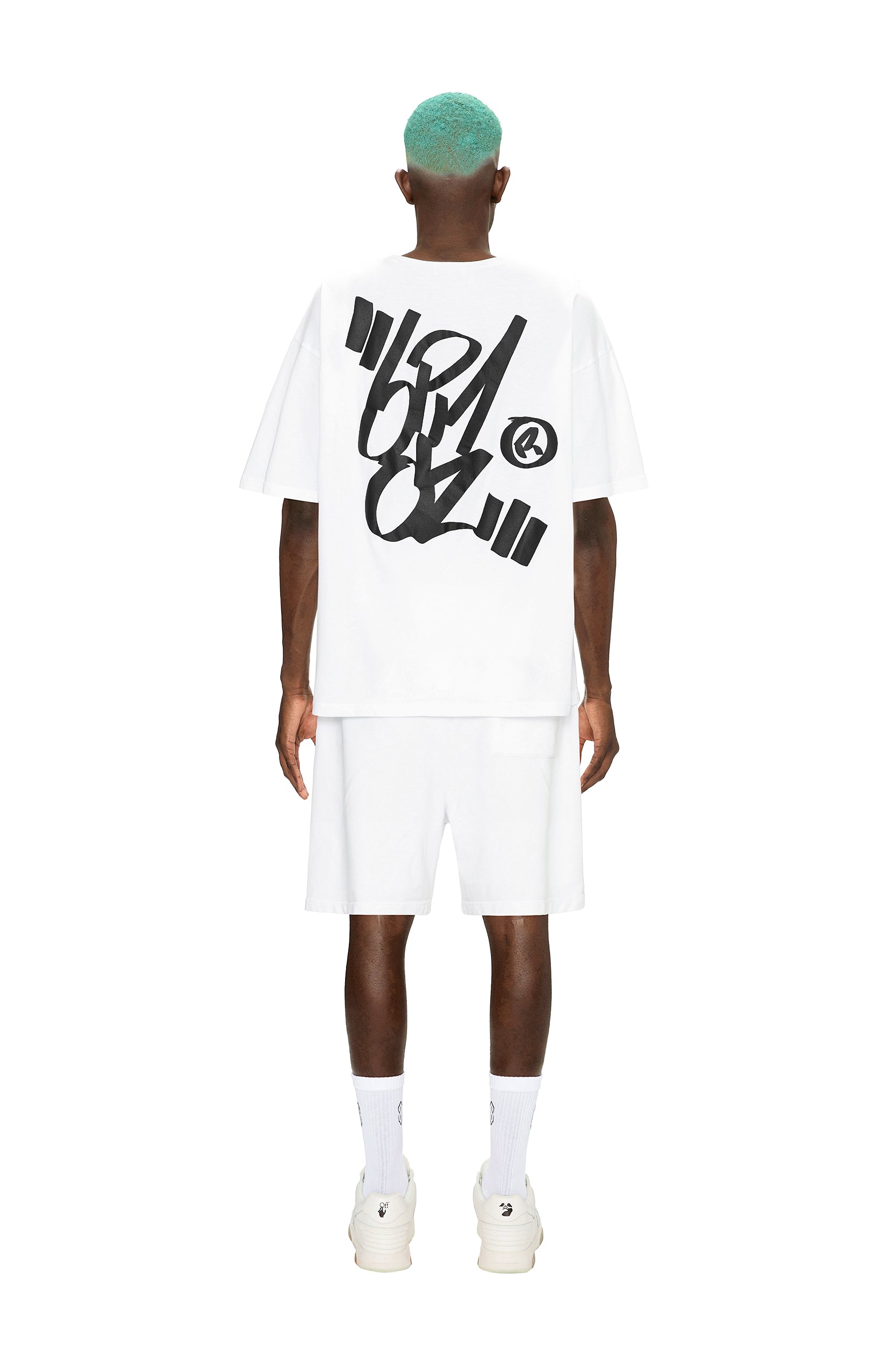 A person with turquoise hair faces away, wearing the 6PM SIGNATURE T-SHIRT WHITE featuring bold black graffiti lettering on the back, paired with white shorts, socks, and sneakers, all set against a plain white background.