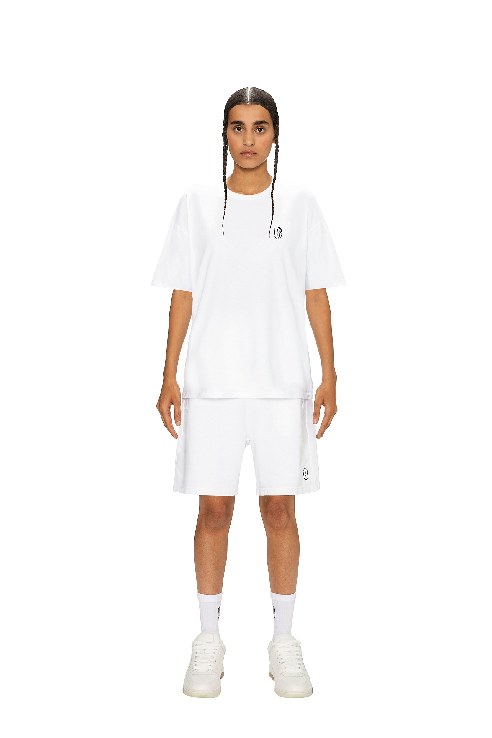 A person with long braids stands facing forward in a 6PM SIGNATURE T-SHIRT WHITE, paired with white shorts, socks, and sneakers against a plain white background.