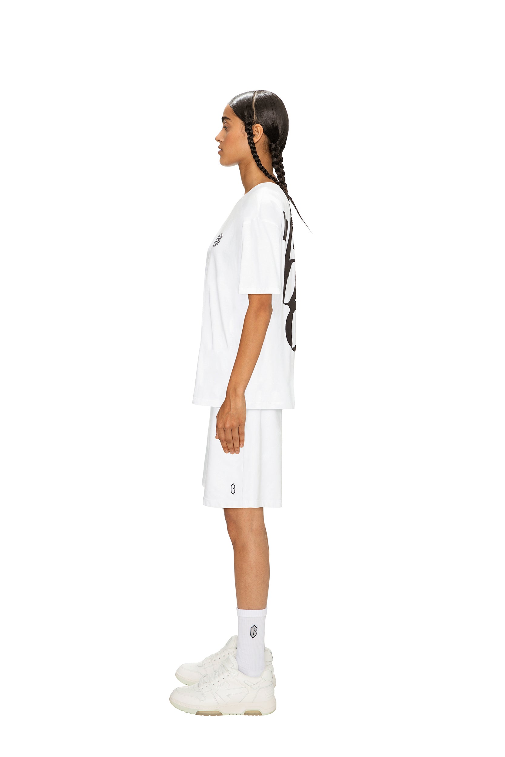 A person with long, dark braided hair stands in profile against a white background, wearing the 6PM SIGNATURE T-SHIRT WHITE, paired with white shorts, crew socks, and sneakers.