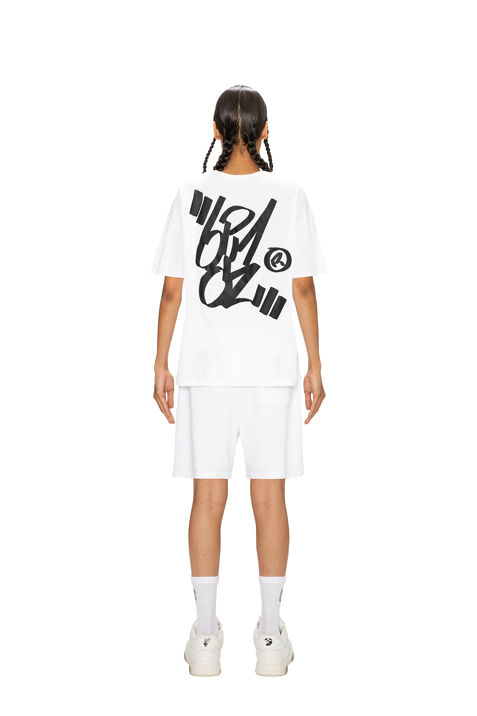 A person with braided hair faces away, wearing the 6PM SIGNATURE T-SHIRT WHITE featuring bold black graffiti-style text on the back, paired with white shorts, socks, and sneakers, all set against a plain white background.