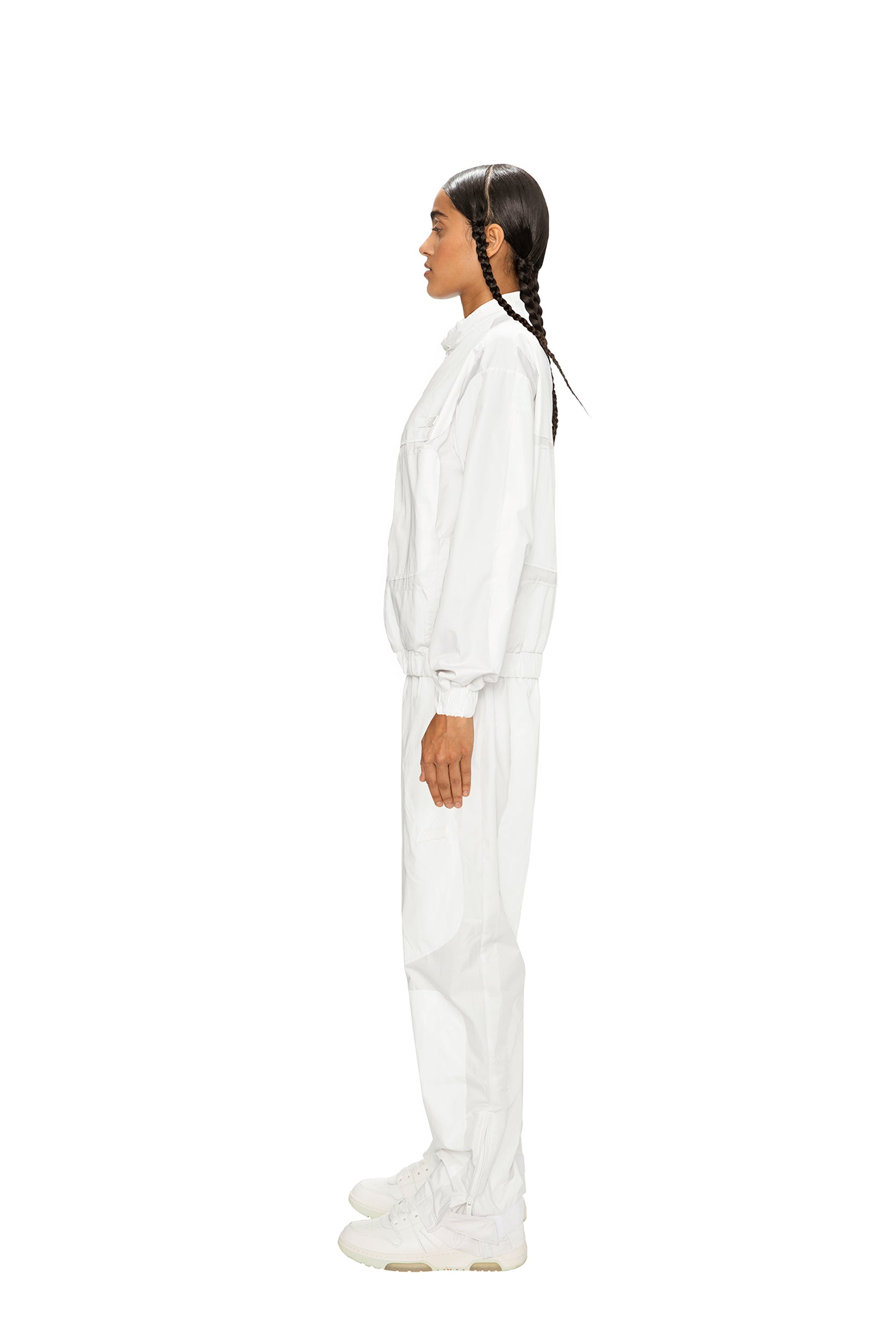 A person with braided hair, wearing 6PM TRACK PANTS WHITE and sneakers, stands in profile against a plain white background.