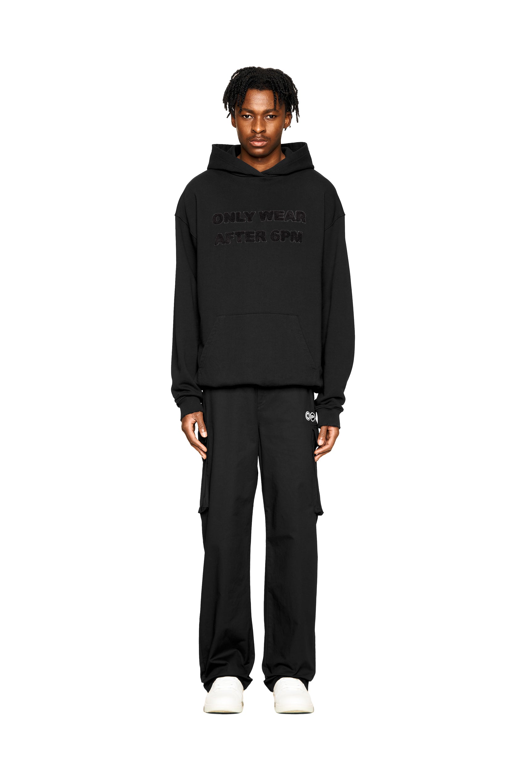 A person stands facing forward against a white background, wearing the 6PM ONLY WEAR AFTER 6PM HOODIE 2.0 in black, black pants, and white slip-on shoes.