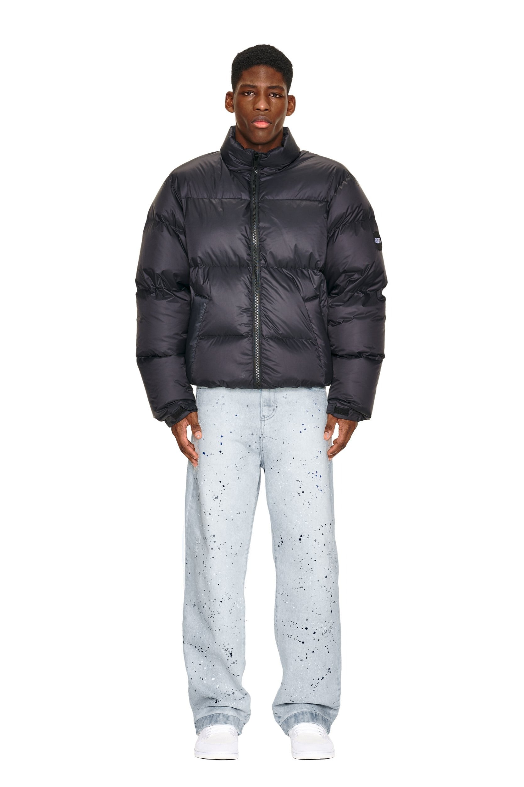 A man stands facing forward wearing the 6PM DOWN JACKET BLACK MATTE, light blue paint-splatter jeans, and white shoes against a plain white background.