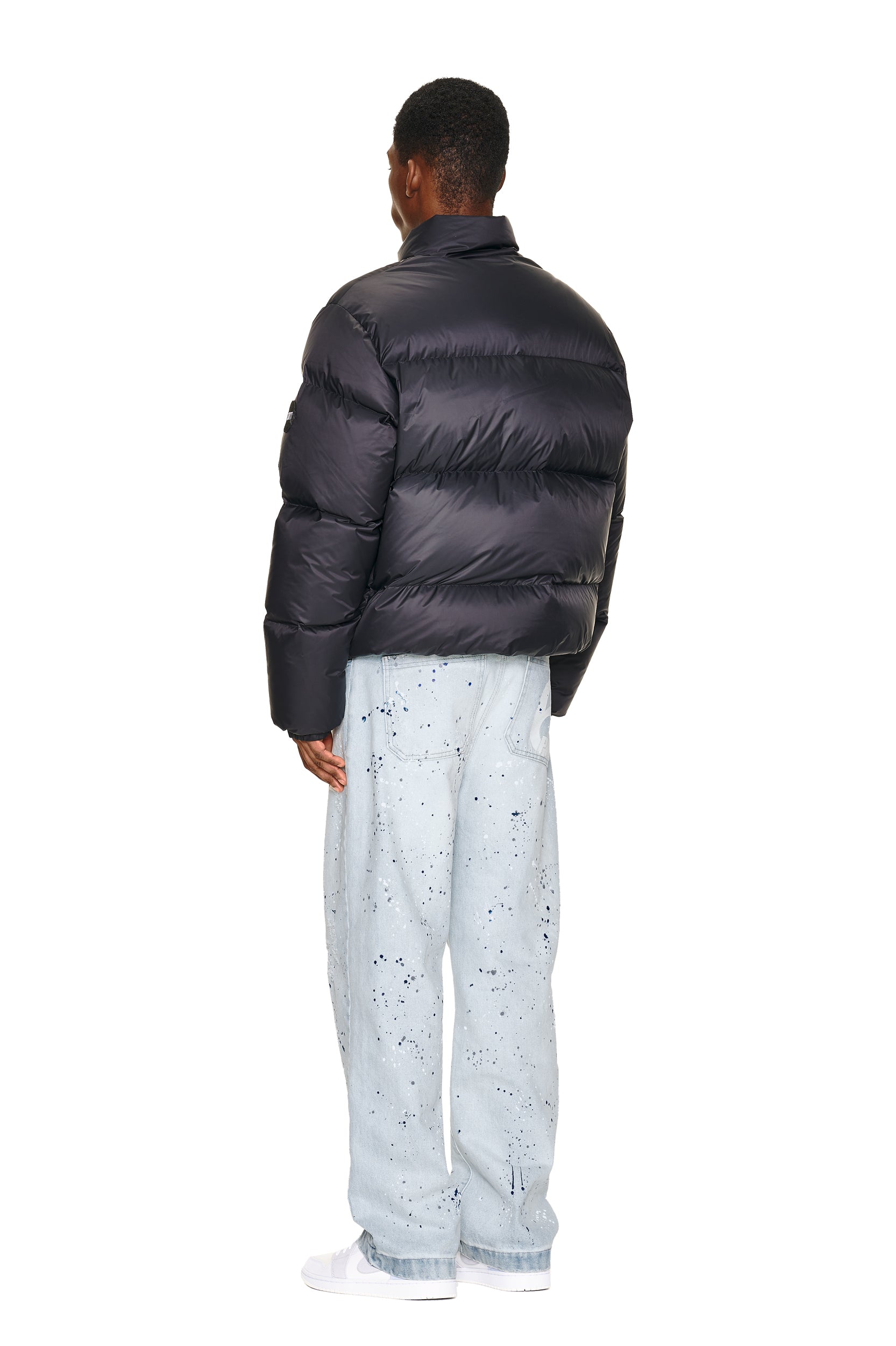 A person stands facing away against a white background, wearing the 6PM DOWN JACKET BLACK MATTE with light blue paint-splattered baggy jeans and white sneakers.