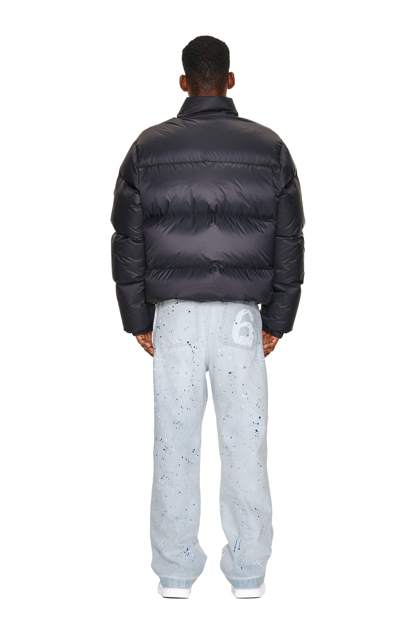 A man faces away, wearing the 6PM DOWN JACKET BLACK MATTE and light blue jeans with blue paint splatters plus a large white "6" on the back pocket. He pairs them with white sneakers, all set against a white background.