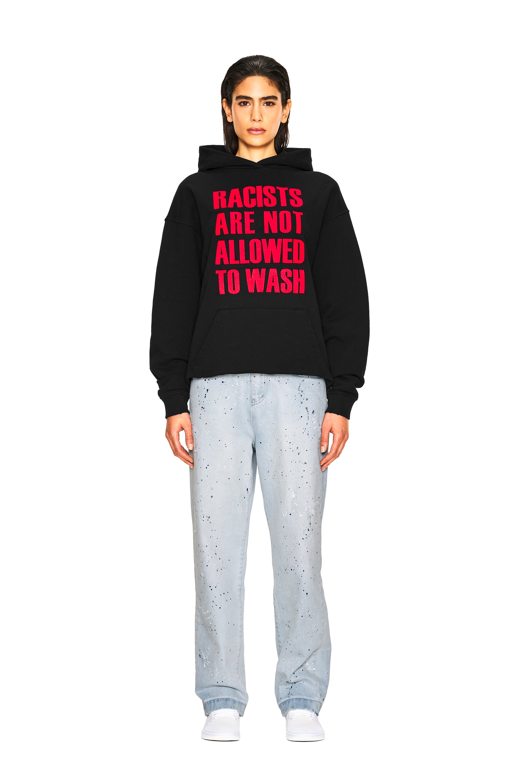 A person stands facing forward in the 6PM NO RACISTS HOODIE BLACK, light blue paint-splattered jeans, and white sneakers against a plain white background.