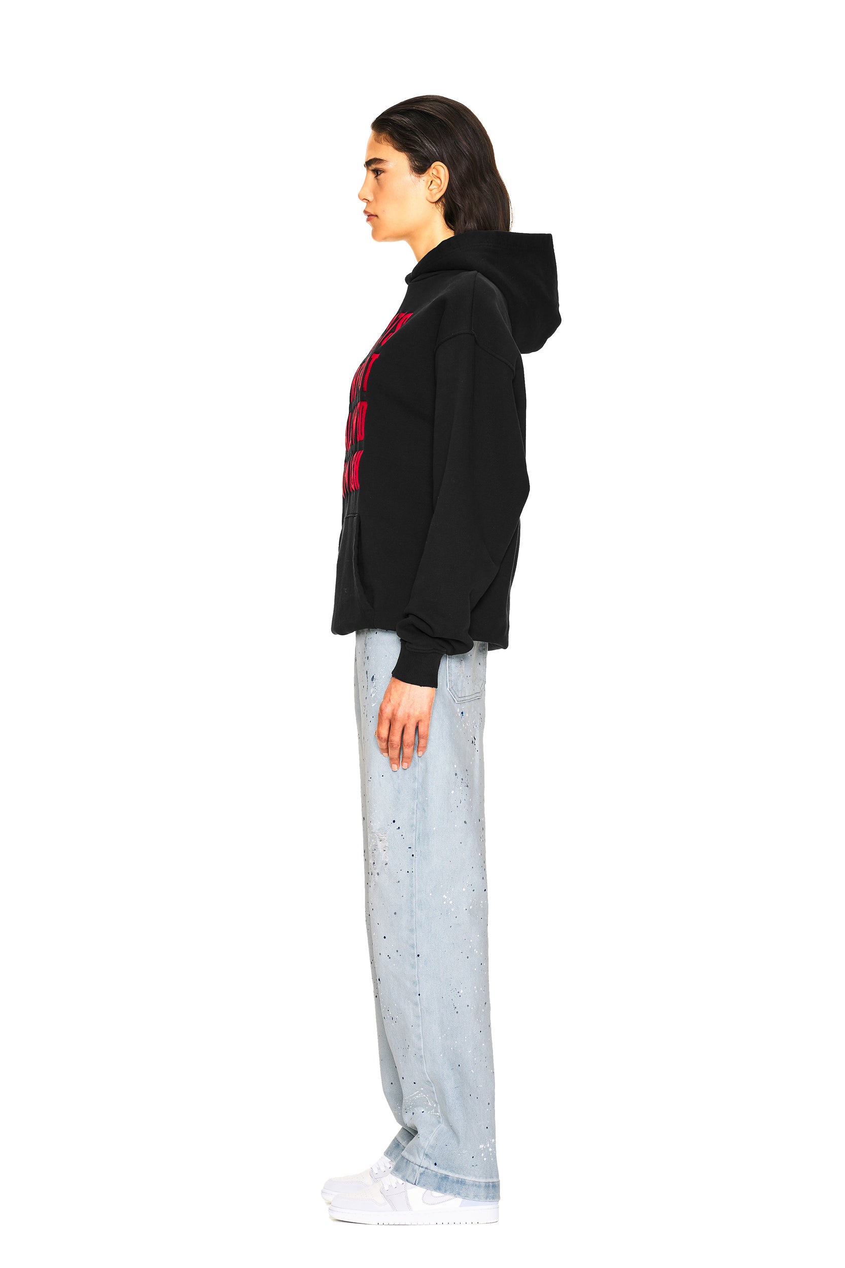 A person stands in profile against a plain white background wearing the 6PM NO RACISTS HOODIE BLACK, light blue speckled jeans, and white sneakers.
