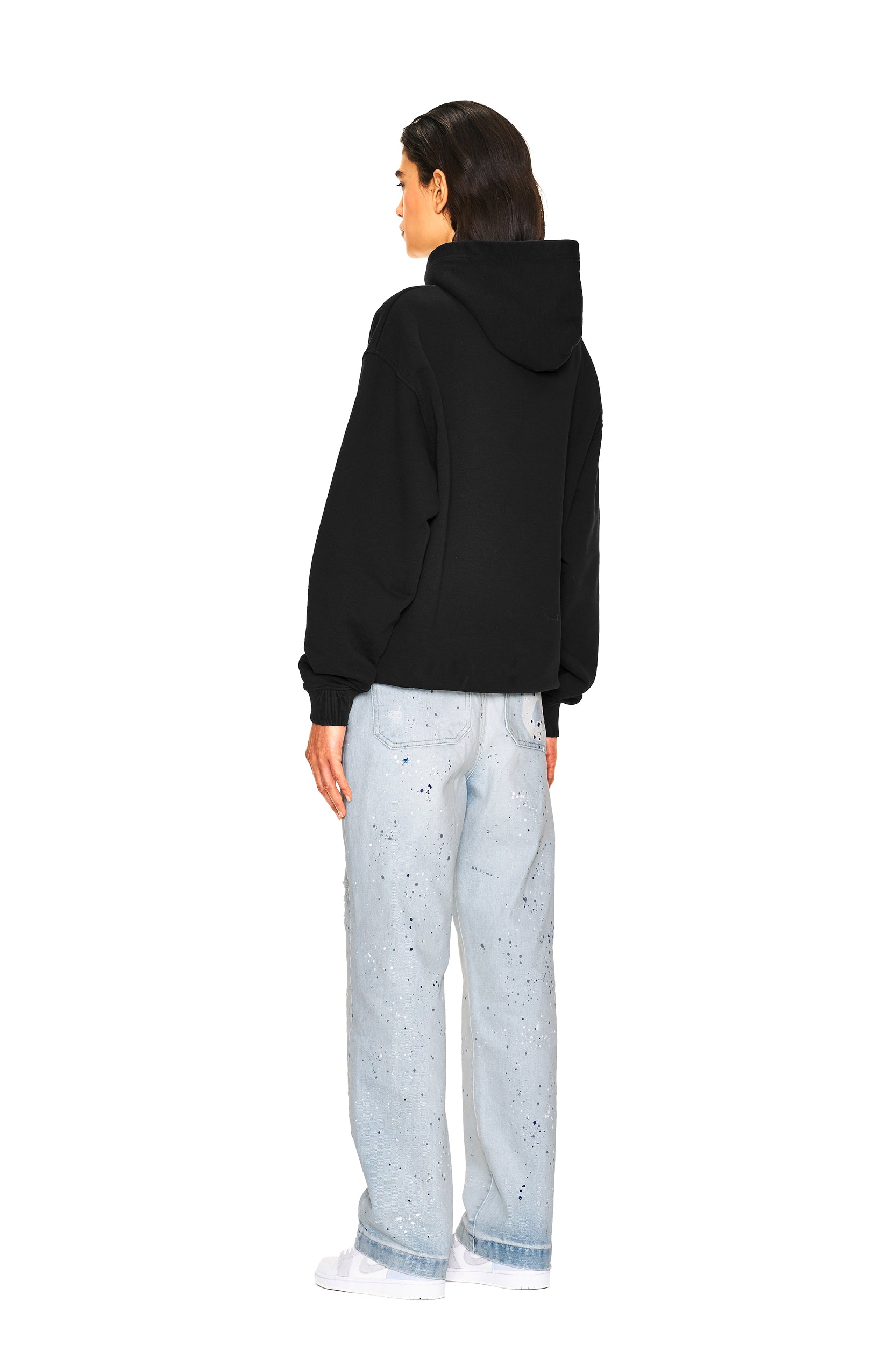 A person stands facing away in a 6PM NO RACISTS HOODIE BLACK with the hood down, paired with light blue, paint-splattered jeans, against a plain white background.