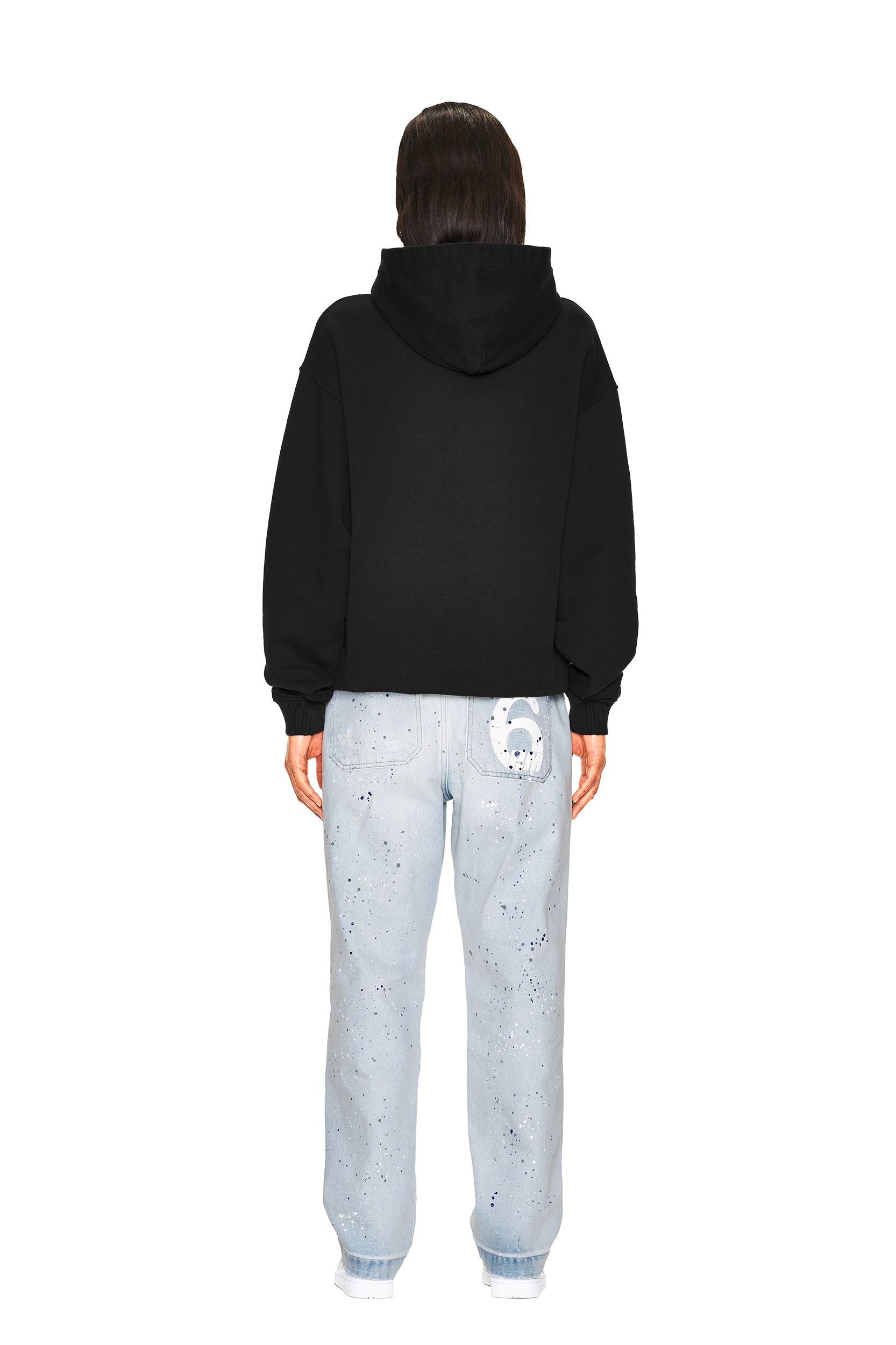 A person faces away in a 6PM NO RACISTS HOODIE BLACK, paired with light blue, paint-splattered jeans featuring a white graphic patch on the back pocket. The backdrop is plain white.