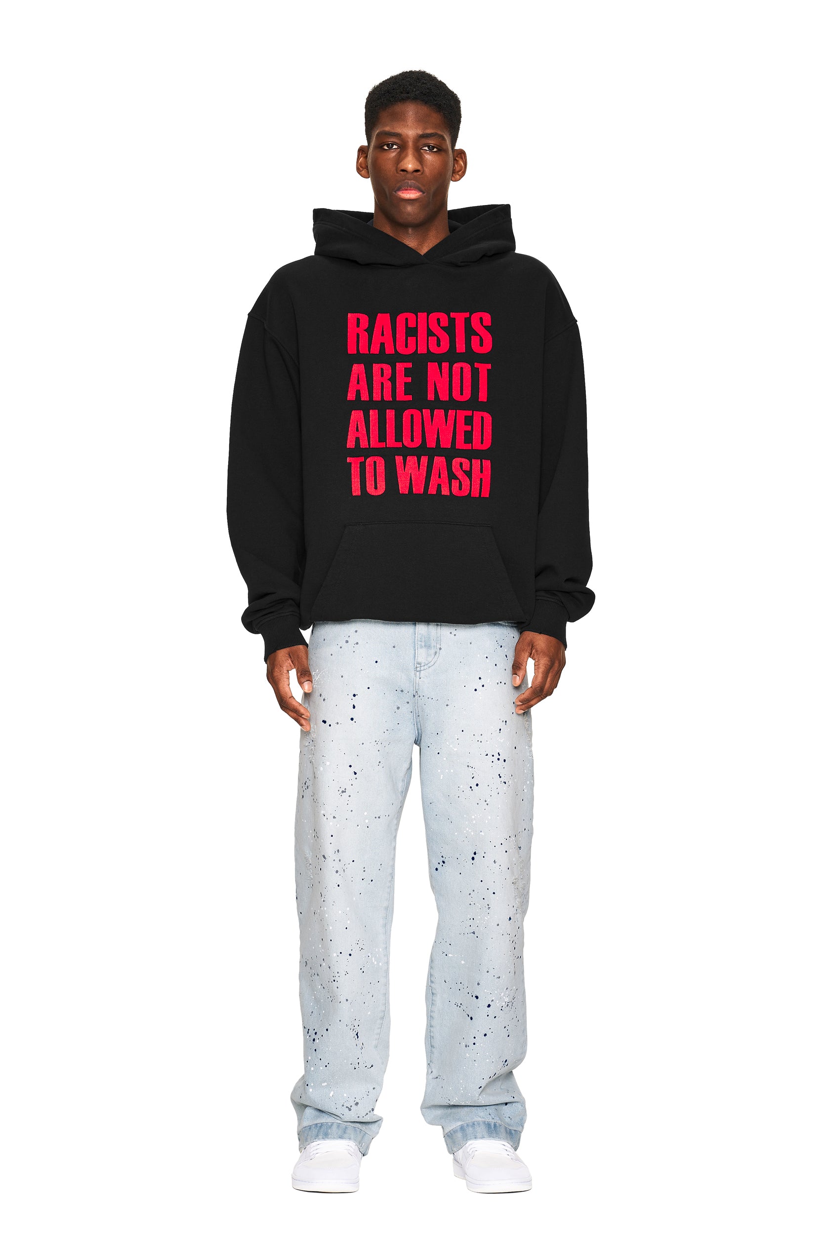 A man faces forward wearing the 6PM NO RACISTS HOODIE BLACK with bold red text, paired with light blue paint-splattered jeans and white sneakers, against a plain white background.