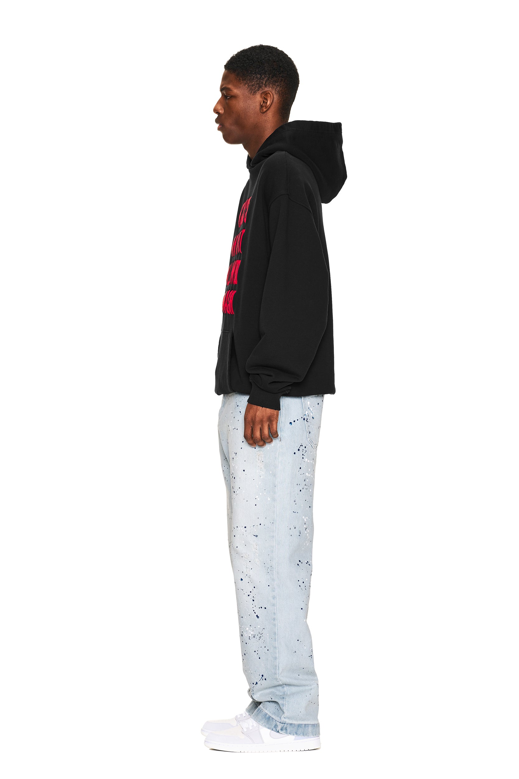A young man stands in profile against a white background, wearing the 6PM NO RACISTS HOODIE BLACK, light blue paint-splattered jeans, and white sneakers.