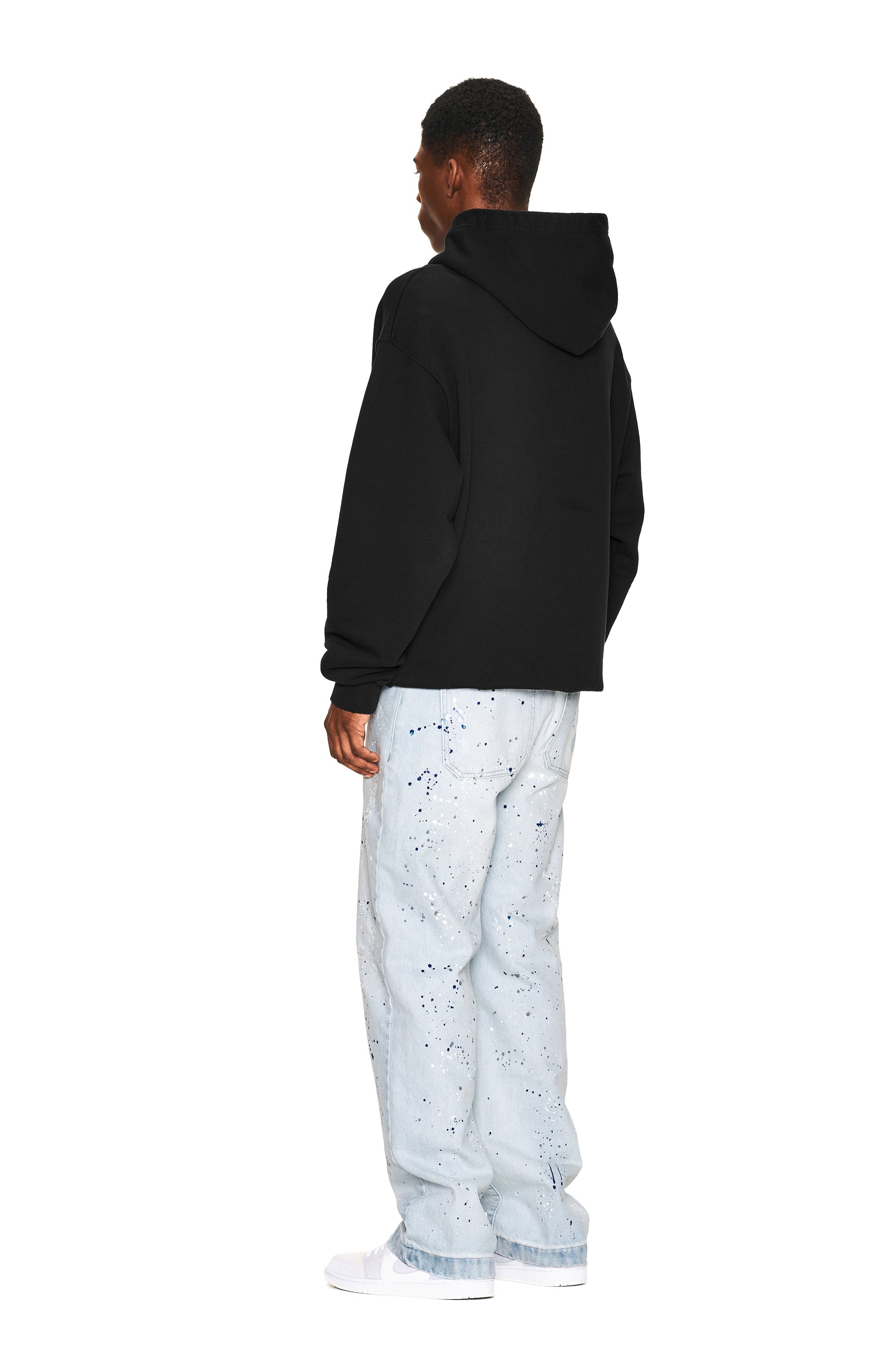 A person stands facing away in a 6PM NO RACISTS HOODIE BLACK, paired with light blue paint-splattered jeans and white sneakers, against a plain white background.