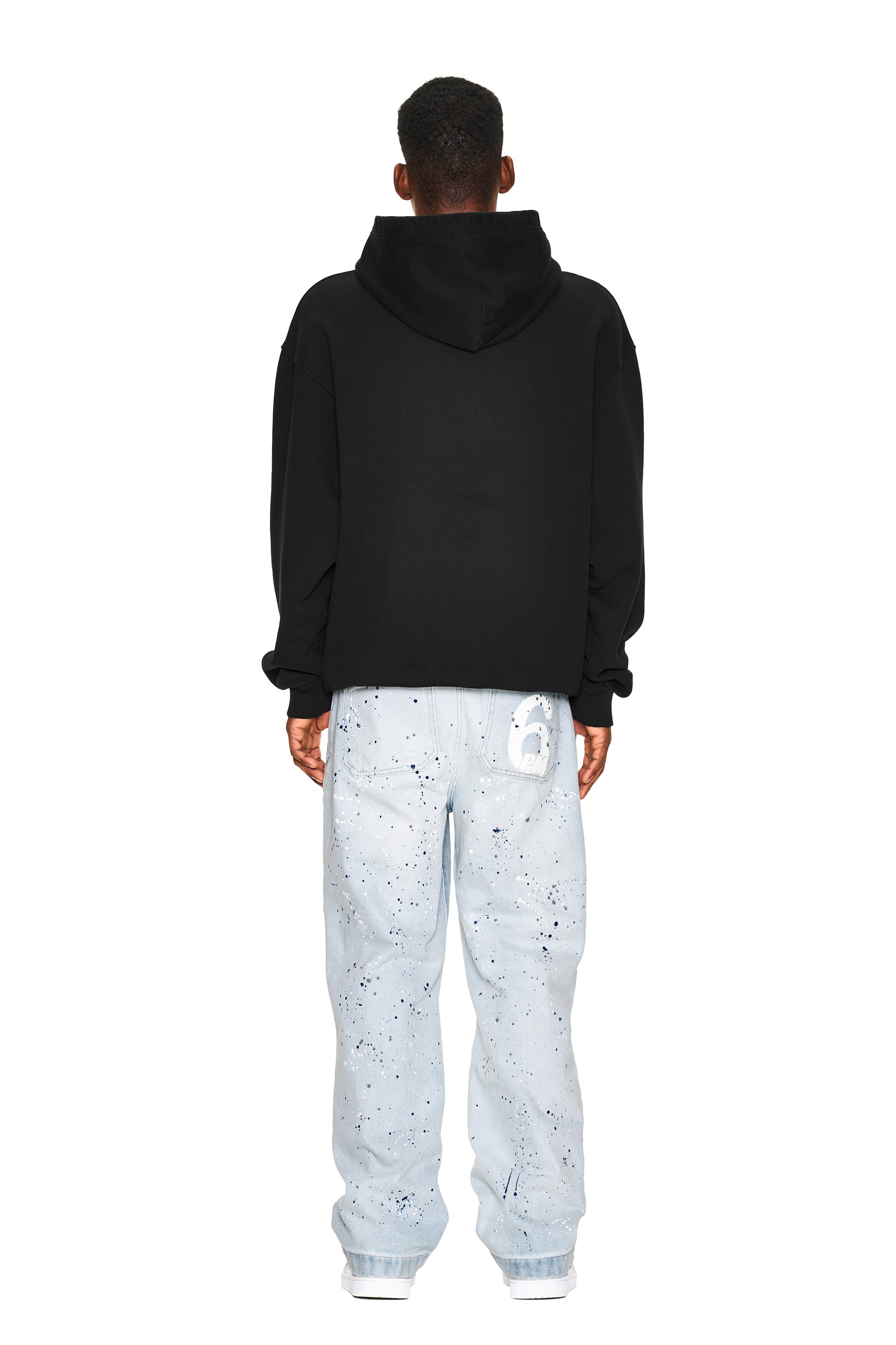 A person faces away, wearing the 6PM NO RACISTS HOODIE BLACK and loose light blue jeans with black paint splatters and a white graphic on the back pocket, plus white sneakers, all set against a plain white background.