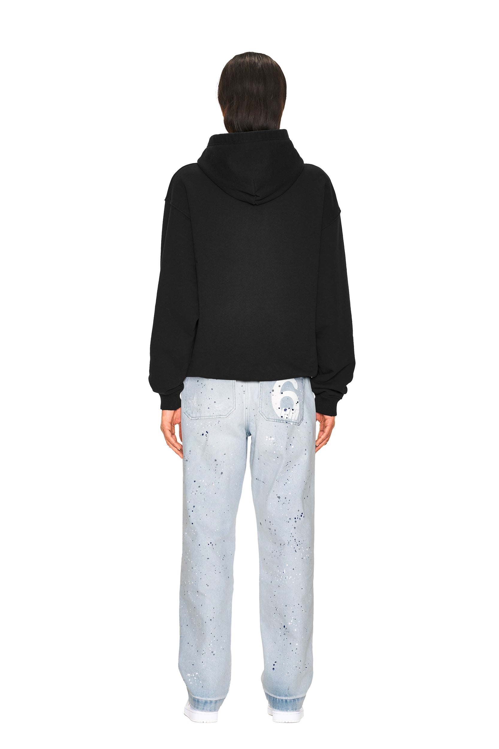 A person faces away, wearing the 6PM LOGO ZIP HOODIE BLACK and light blue jeans with paint splatters and a cartoon face patch on the back pocket, against a plain white background.