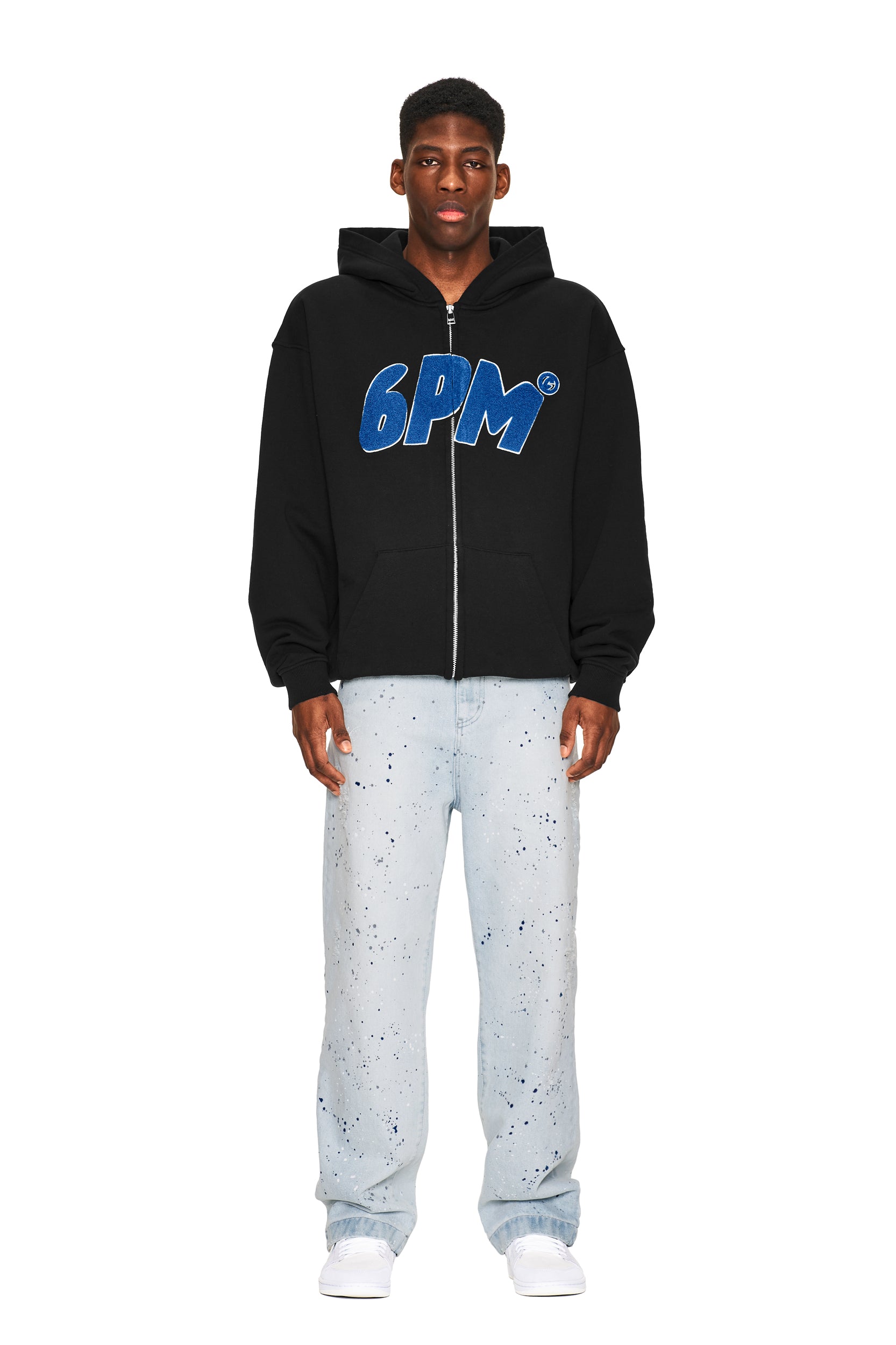 A man stands facing forward in a 6PM LOGO ZIP HOODIE BLACK, light blue paint-splatter jeans, and white sneakers against a plain white background.
