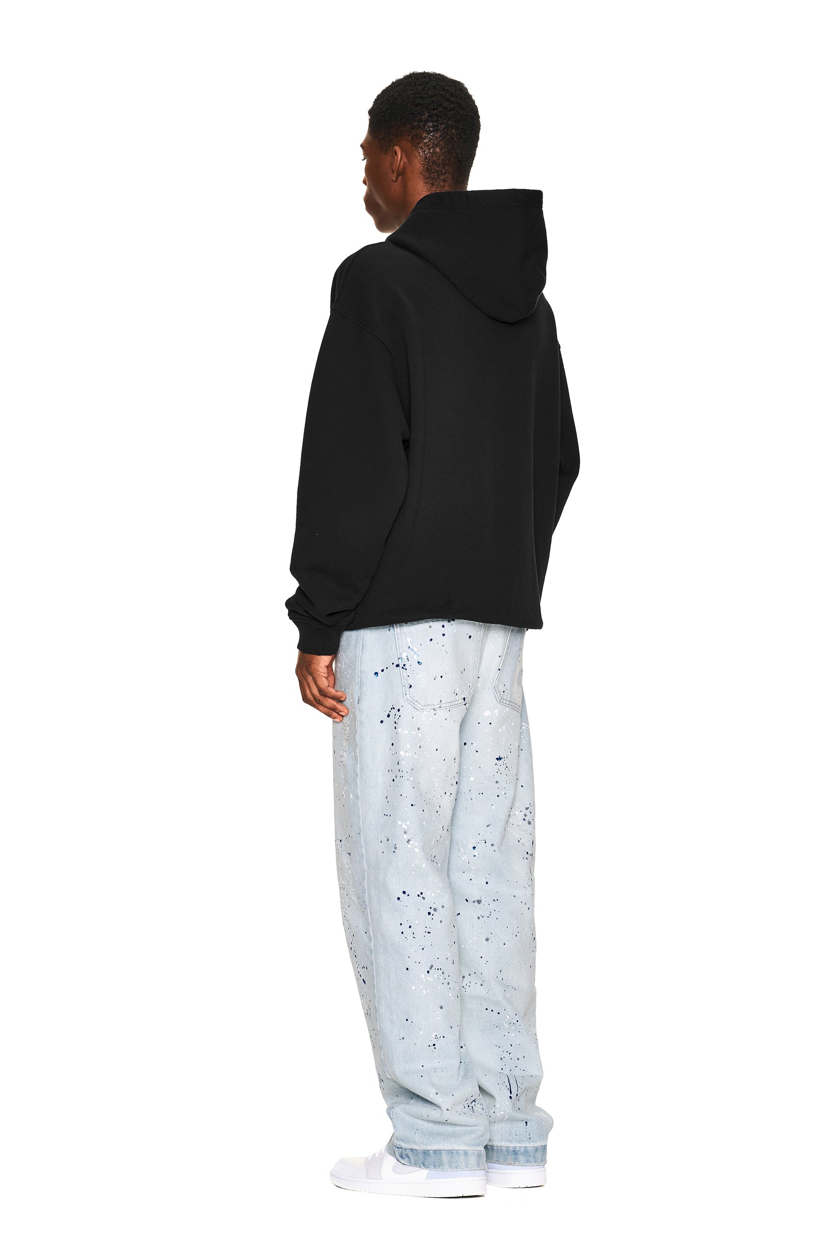 A person stands facing away against a white background, wearing the 6PM LOGO ZIP HOODIE BLACK, light blue paint-splattered jeans, and white sneakers.