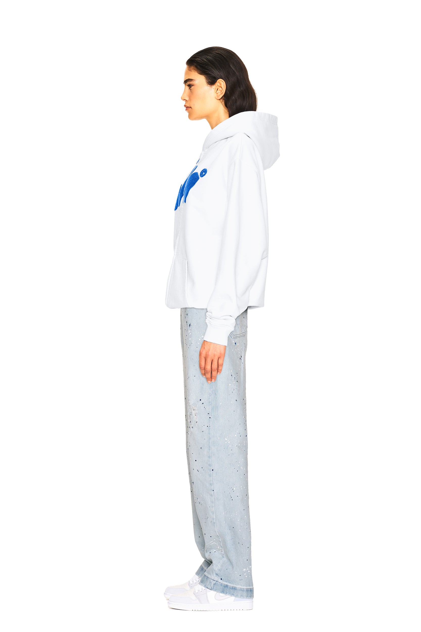 A woman stands in profile wearing JEANS PANTS BLUE by 6PM, styled with a white hoodie featuring a blue design and white sneakers, set against a plain white background.