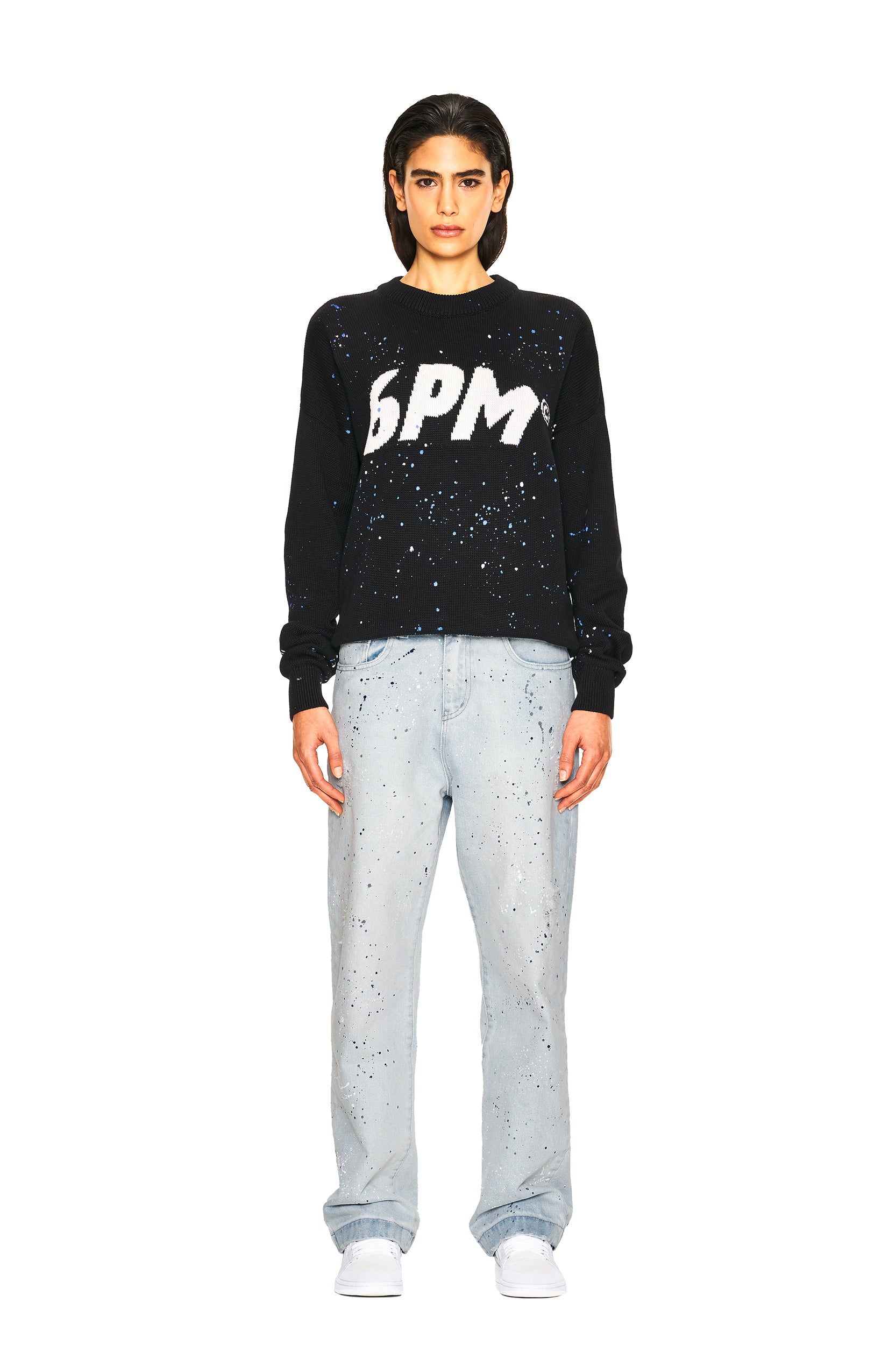 A person stands facing forward, wearing the 6PM LOGO KNIT BLACK sweater with large white BPM letters, light blue paint-splattered jeans, and white sneakers against a plain white background.