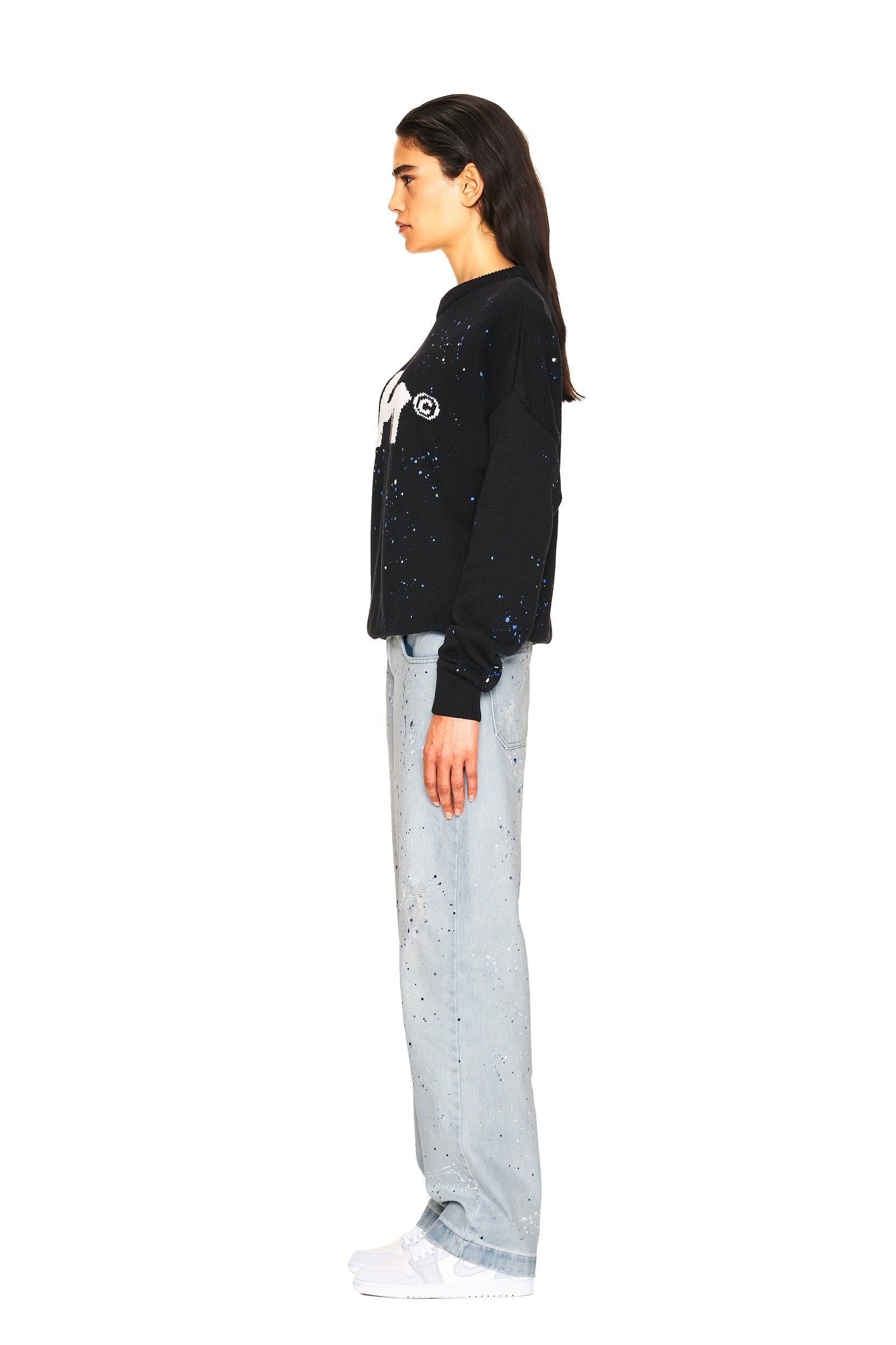 A woman with long dark hair stands in profile wearing the 6PM LOGO KNIT BLACK sweatshirt, light blue speckled jeans, and white sneakers against a plain white background.