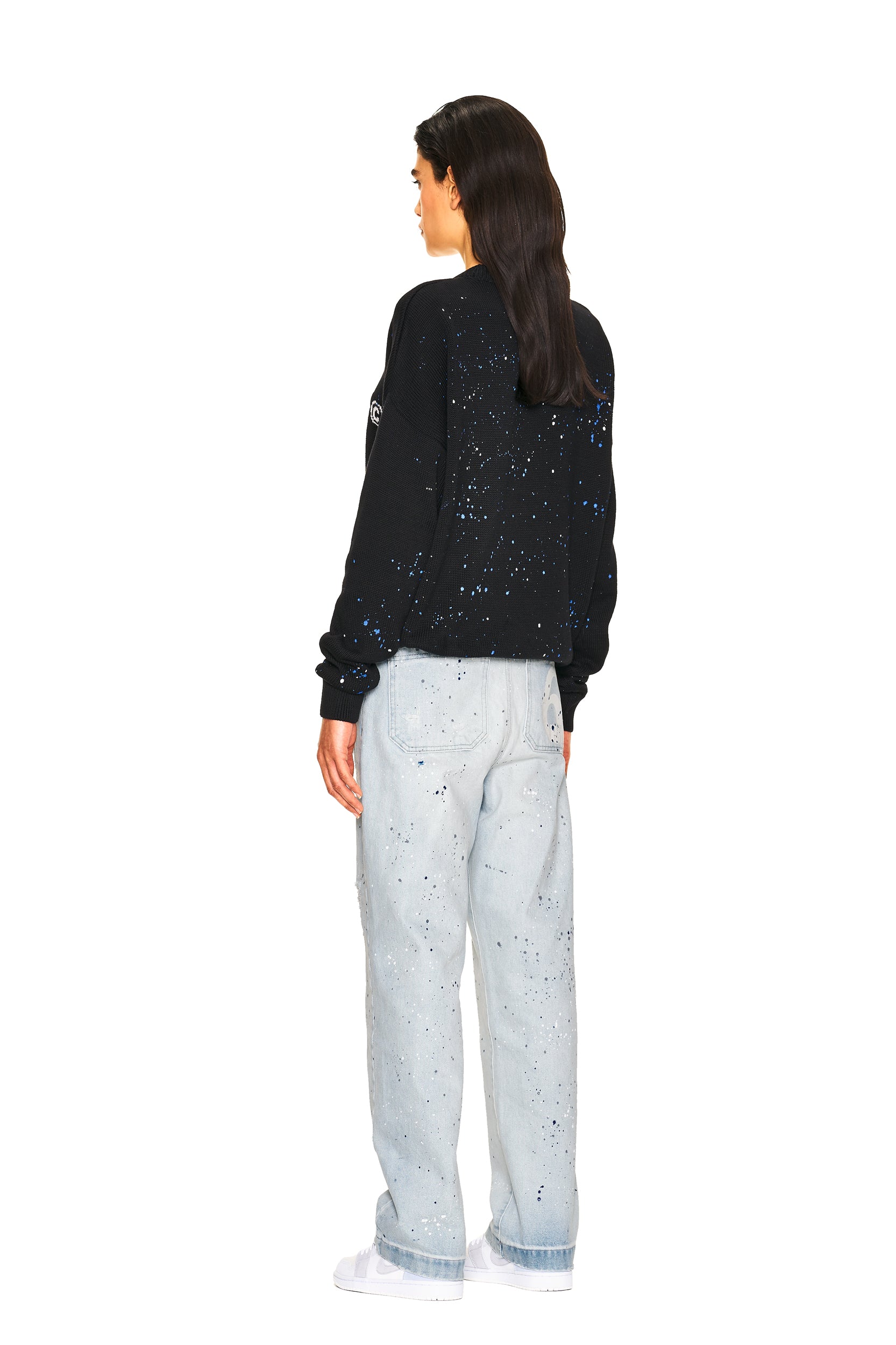 A woman stands facing away in a 6PM LOGO KNIT BLACK sweatshirt with white and blue splatter design, paired with light-wash jeans featuring a matching splatter pattern and white sneakers, set against a white background.