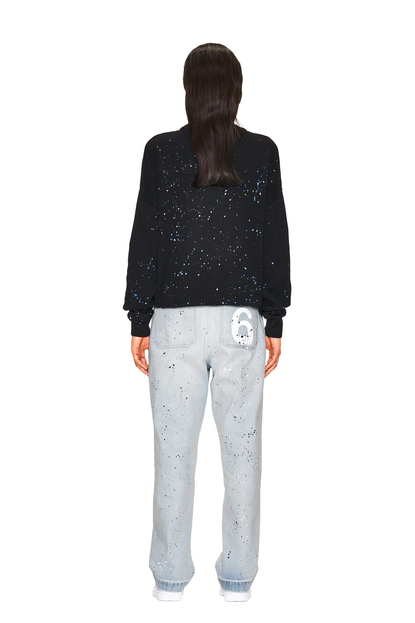 A person stands facing away, wearing the 6PM LOGO KNIT BLACK sweater with a splatter pattern, paired with light blue splatter jeans featuring a cartoon ghost on the back pocket. The background is white.