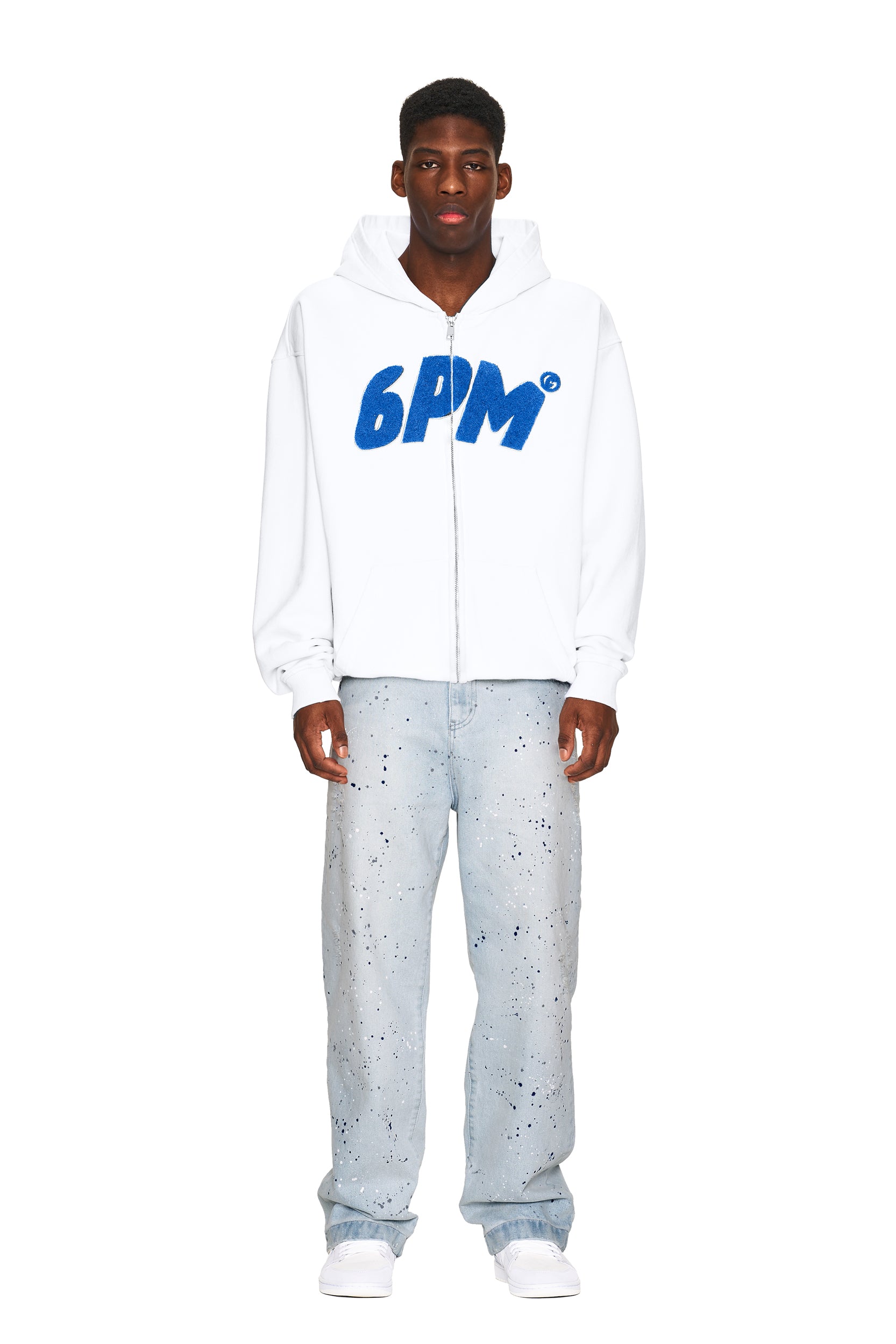 A man faces forward wearing the 6PM LOGO ZIP HOODIE WHITE, featuring bold blue "6PM" lettering, paired with light blue paint-splattered jeans and white sneakers against a plain white background.