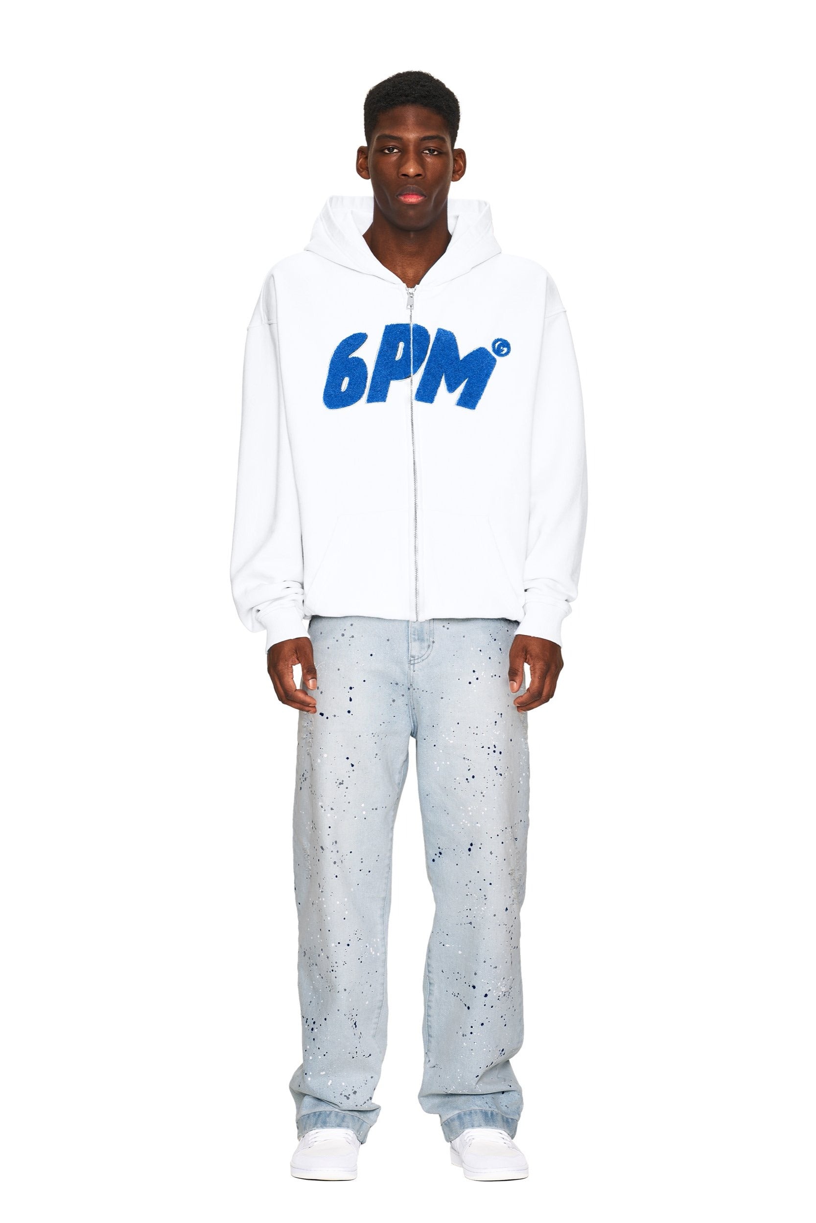 A man faces forward against a plain white background, wearing 6PM’s white zip-up hoodie, JEANS PANTS BLUE from 6PM with a light blue paint-splatter design, and white sneakers.